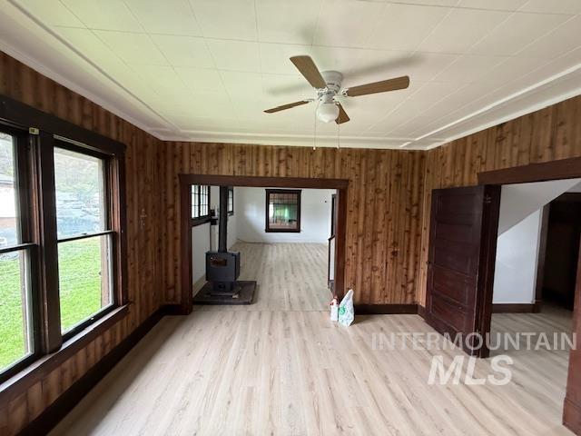 Unfurnished living room with wooden walls, a ceiling fan, a wood stove, light wood-style floors, and ornamental molding