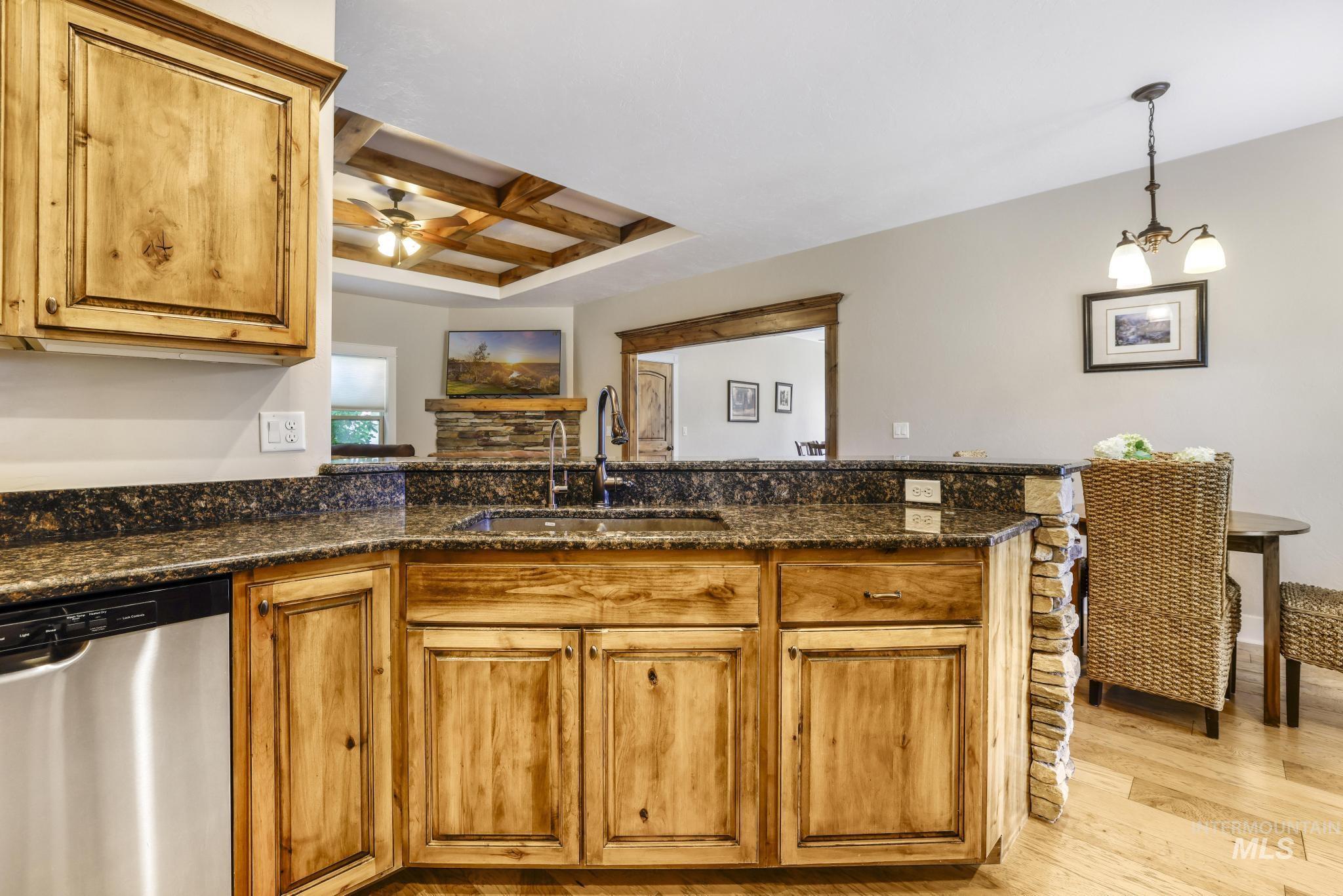 Kitchen featuring stainless steel dishwasher, coffered ceiling, brown cabinetry, hanging light fixtures, and dark stone counters