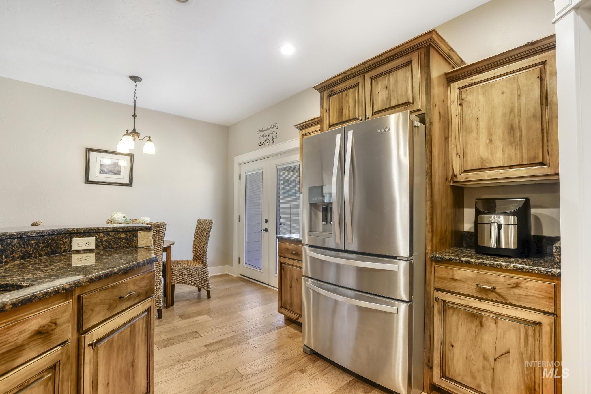 Kitchen with stainless steel refrigerator with ice dispenser, dark stone counters, brown cabinets, hanging light fixtures, and light wood-type flooring