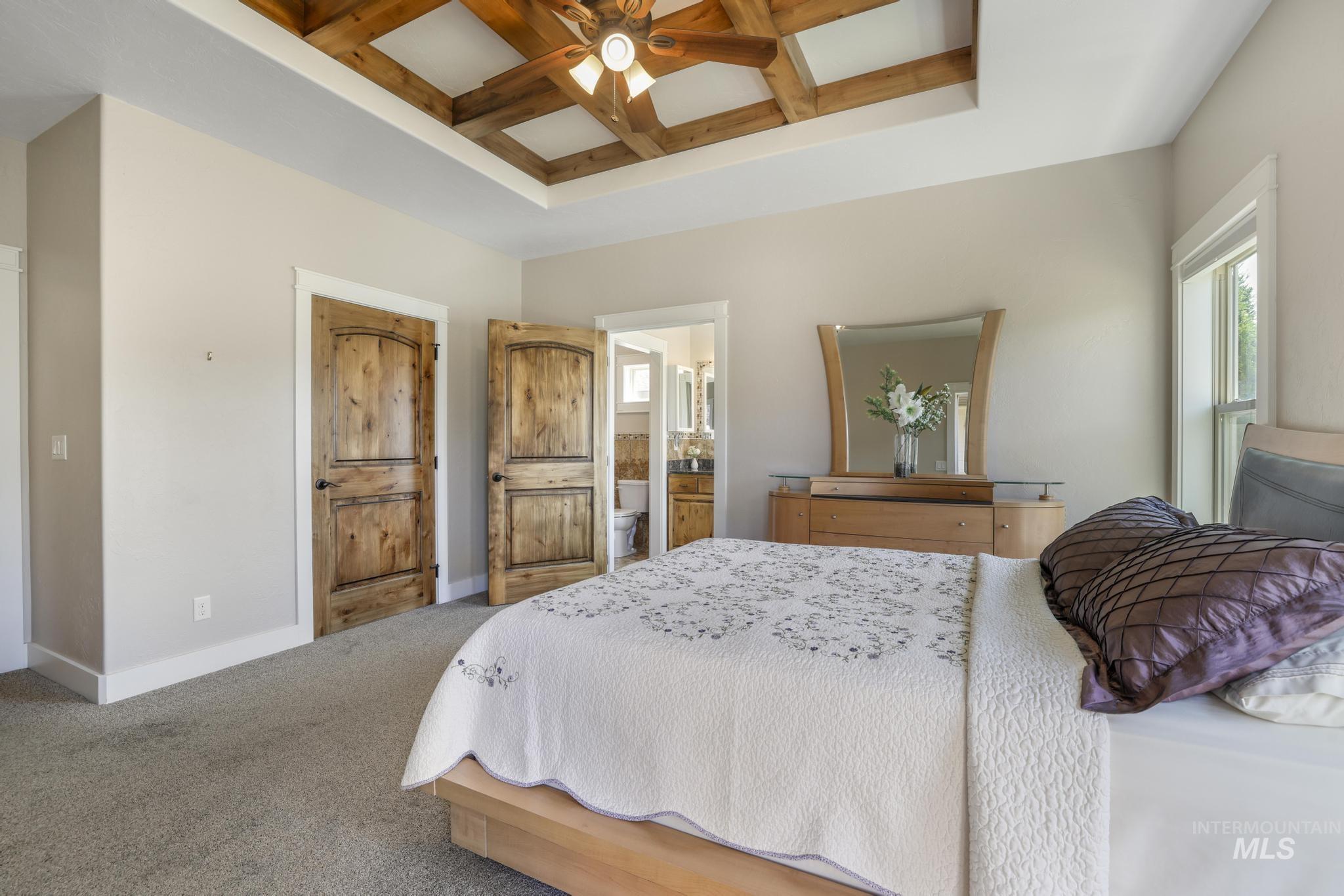 Carpeted bedroom featuring coffered ceiling, beamed ceiling, multiple windows, ceiling fan, and ensuite bath