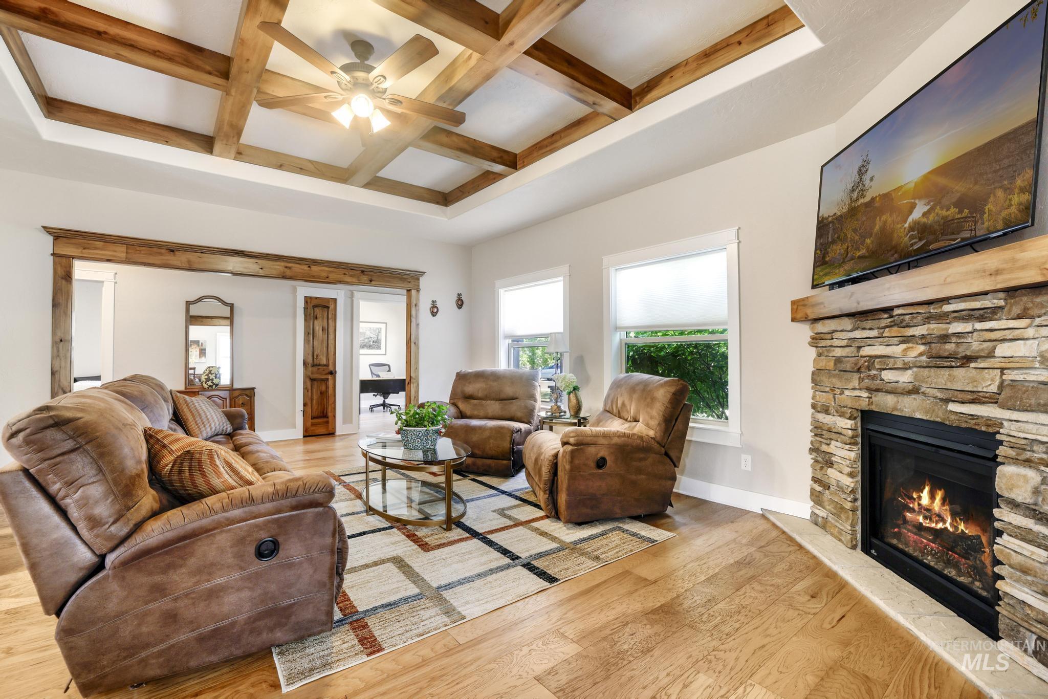 Living area featuring coffered ceiling, beamed ceiling, light wood-style flooring, a stone fireplace, and a ceiling fan