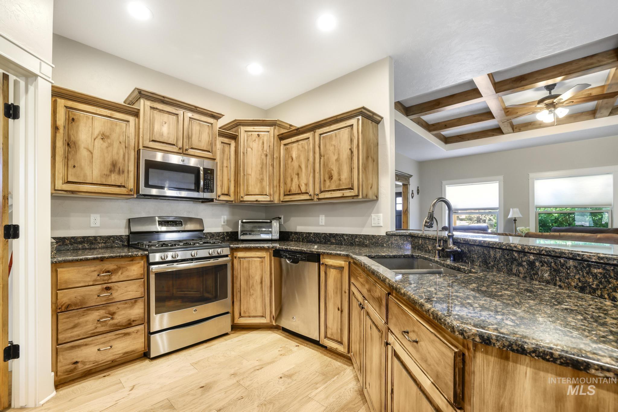Kitchen featuring coffered ceiling, stainless steel appliances, dark stone countertops, beamed ceiling, and light wood finished floors