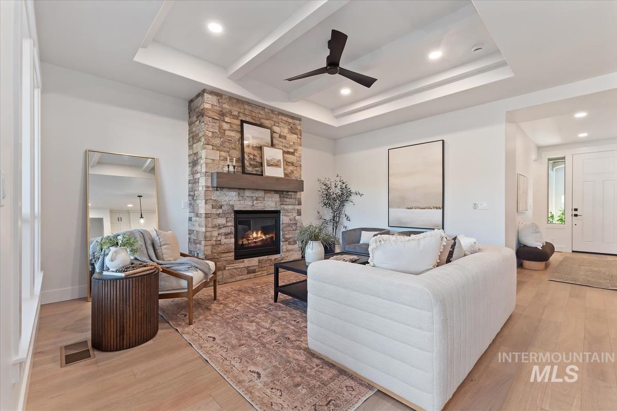 Living area with a raised ceiling, light wood-style flooring, a ceiling fan, a stone fireplace, and recessed lighting