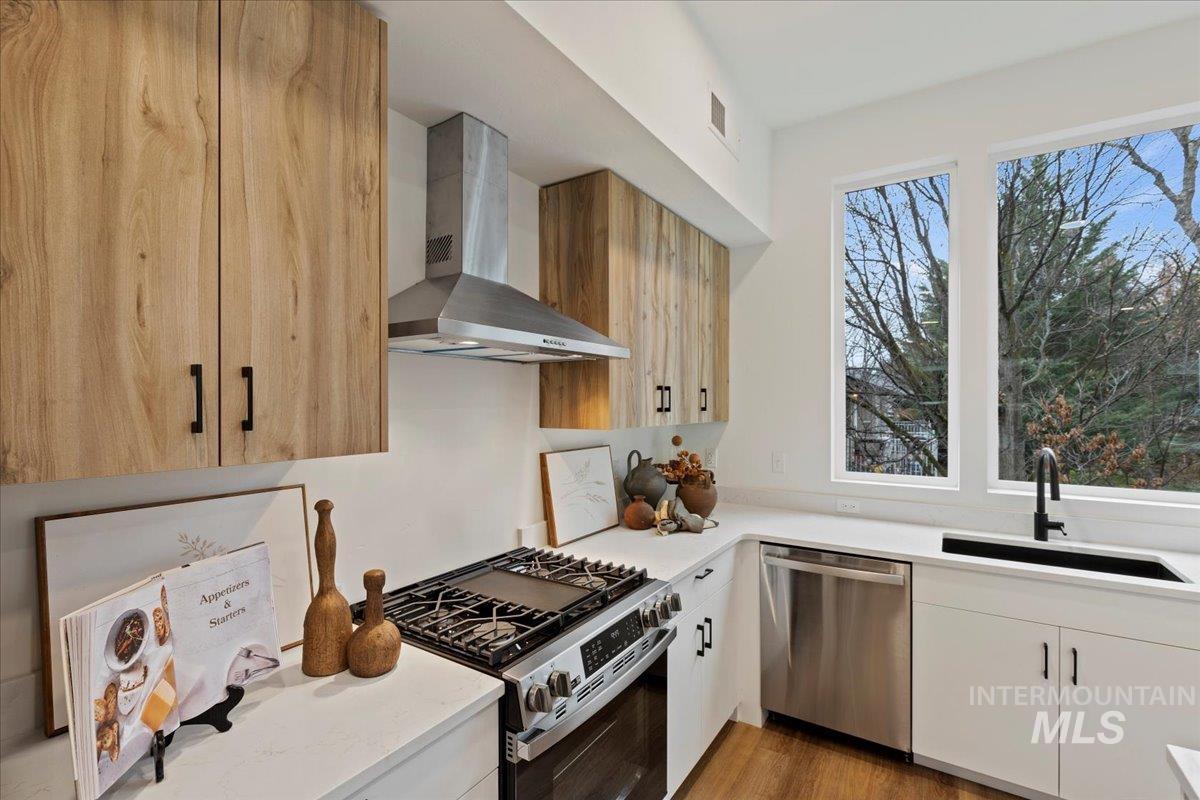 Kitchen with appliances with stainless steel finishes, wall chimney exhaust hood, light stone counters, and light wood-style floors