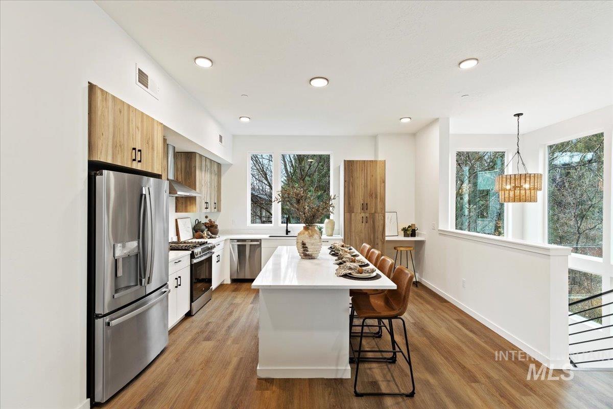 Kitchen featuring stainless steel appliances, a breakfast bar area, hanging light fixtures, wall chimney range hood, and light wood-style flooring