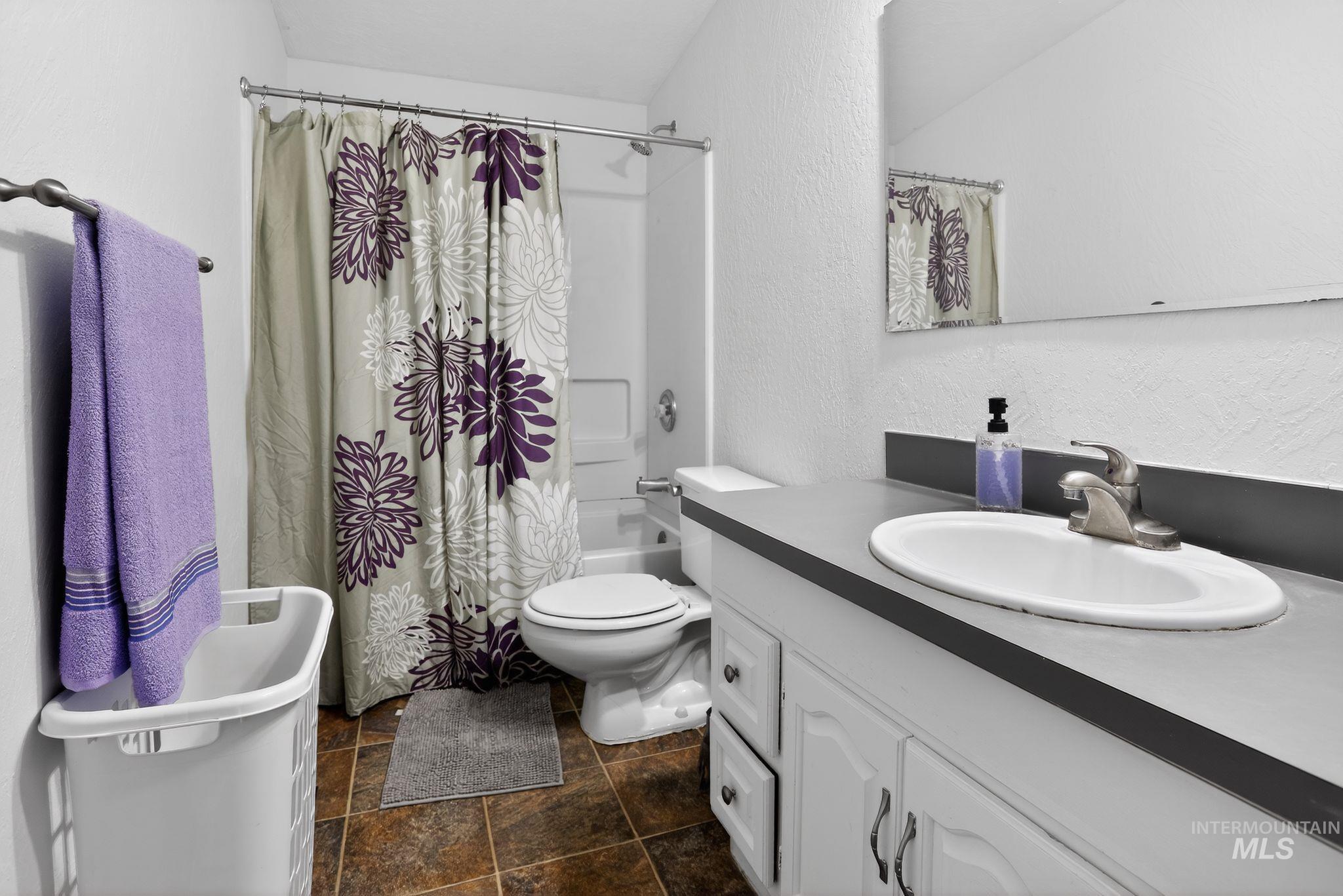 Bathroom featuring a textured wall, vanity, shower / bathtub combination with curtain, and stone tile floors