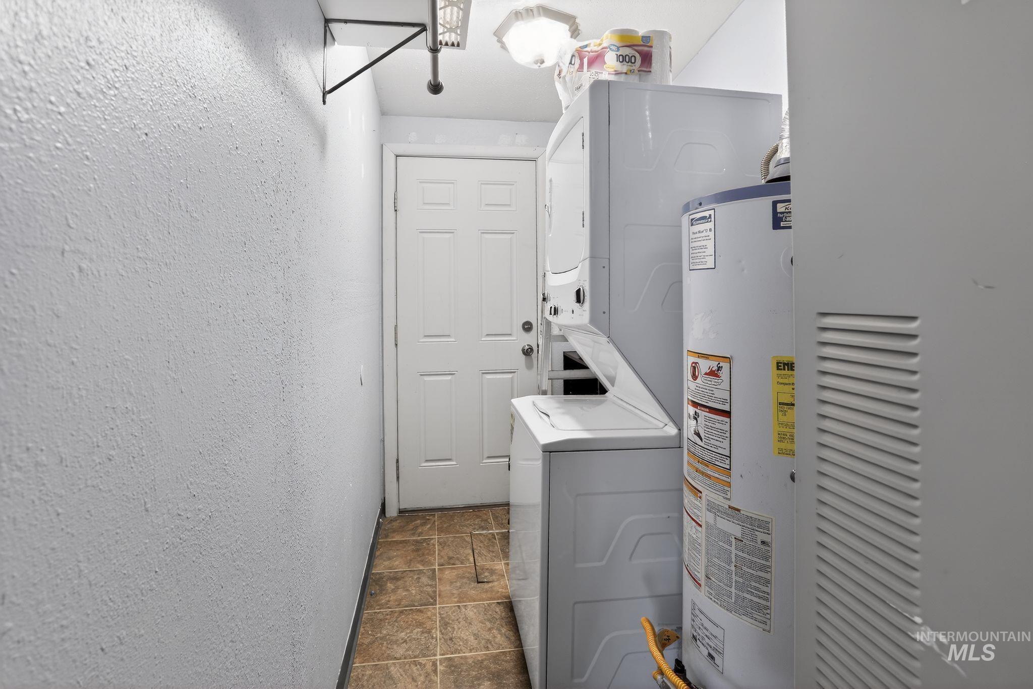 Laundry room with estacked washer and dryer, a textured wall, gas water heater, and stone finish floors