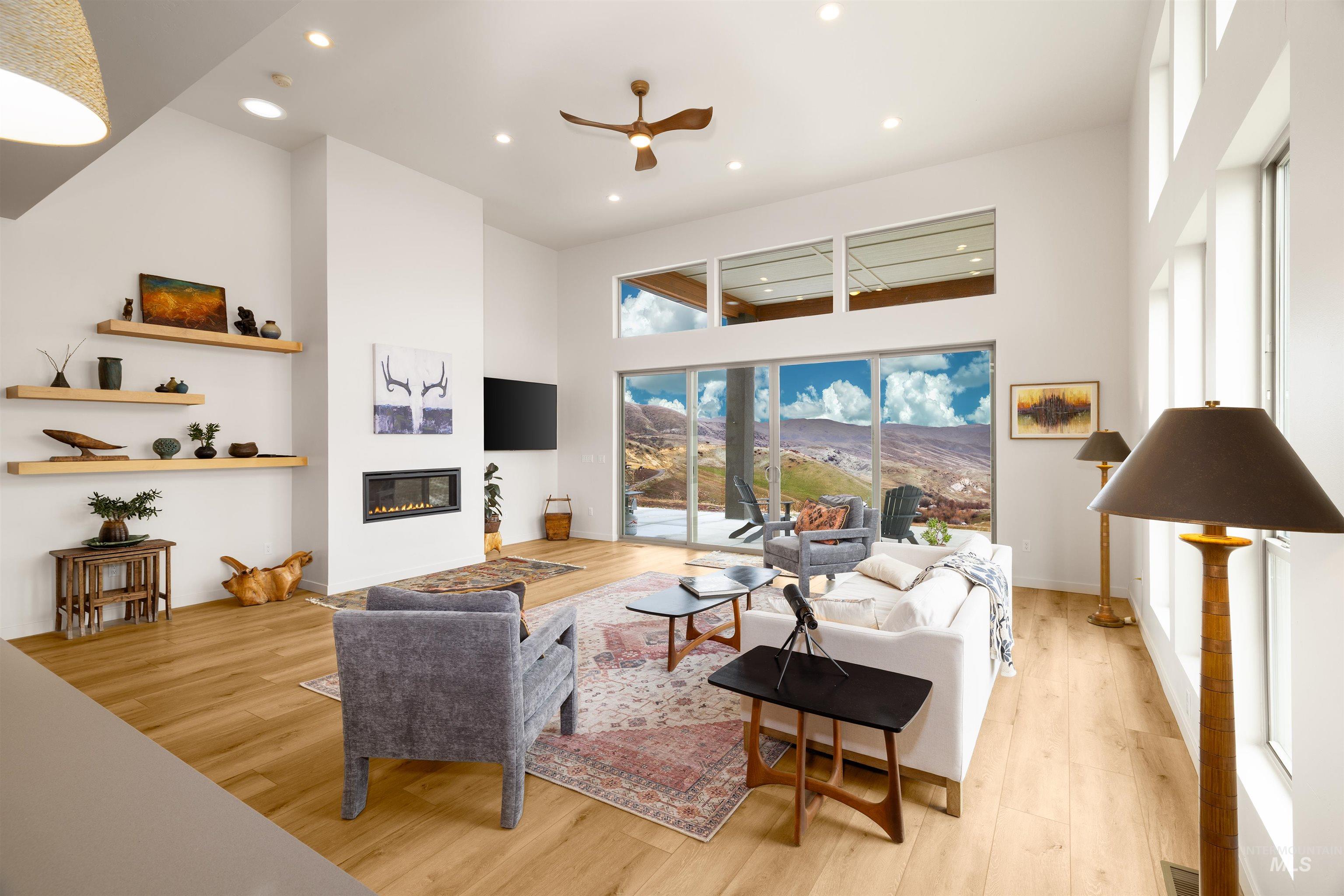 Living area featuring a towering ceiling, light wood finished floors, a glass covered fireplace, ceiling fan, and recessed lighting