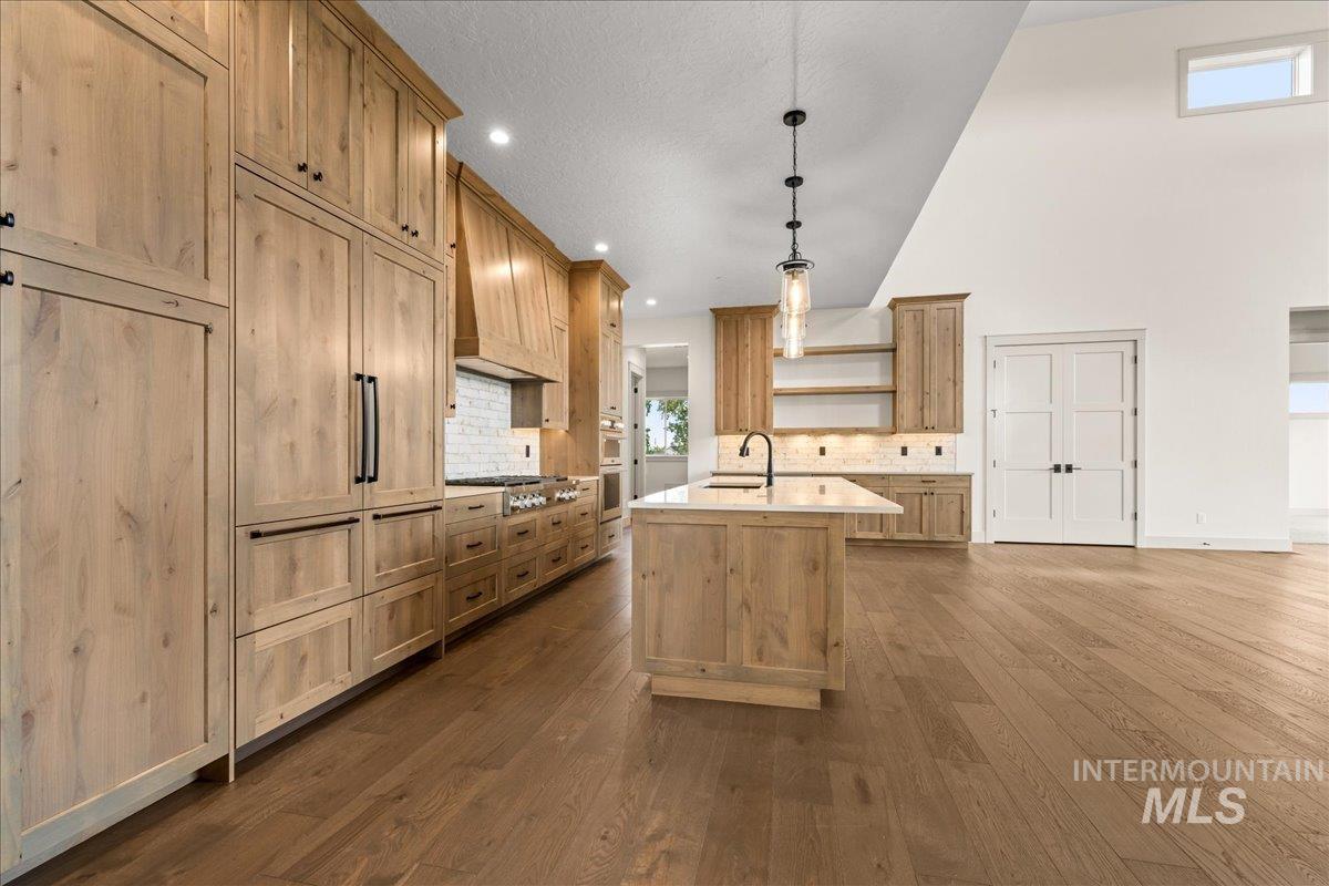 Kitchen featuring open shelves, tasteful backsplash, an island with sink, hanging light fixtures, and dark wood-style floors