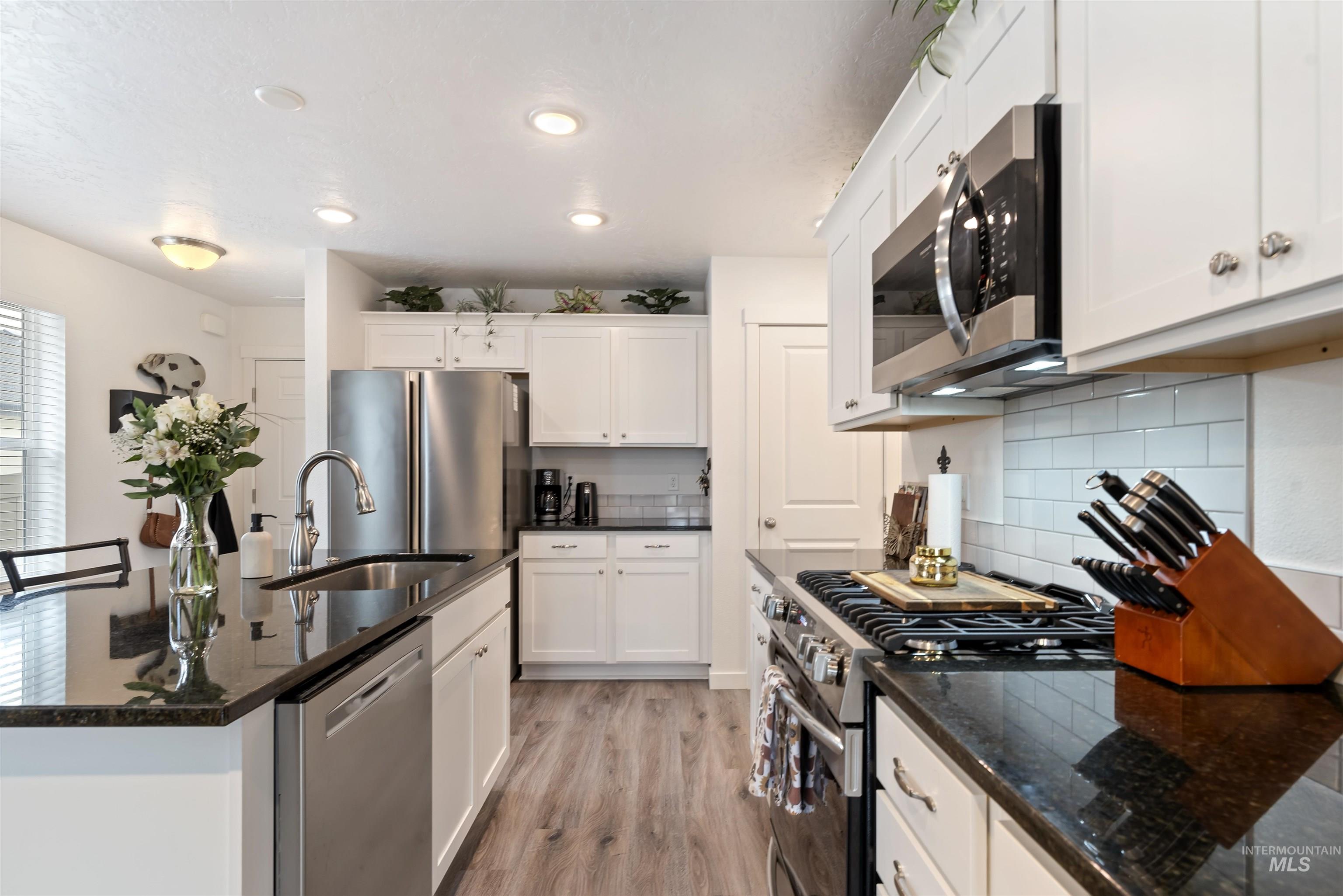 Kitchen with dark stone countertops, appliances with stainless steel finishes, white cabinets, light wood-style flooring, and backsplash