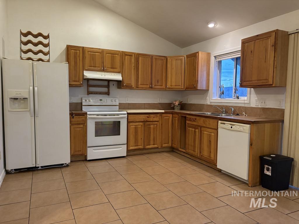 Kitchen with white appliances, under cabinet range hood, brown cabinets, light tile patterned floors, and high vaulted ceiling