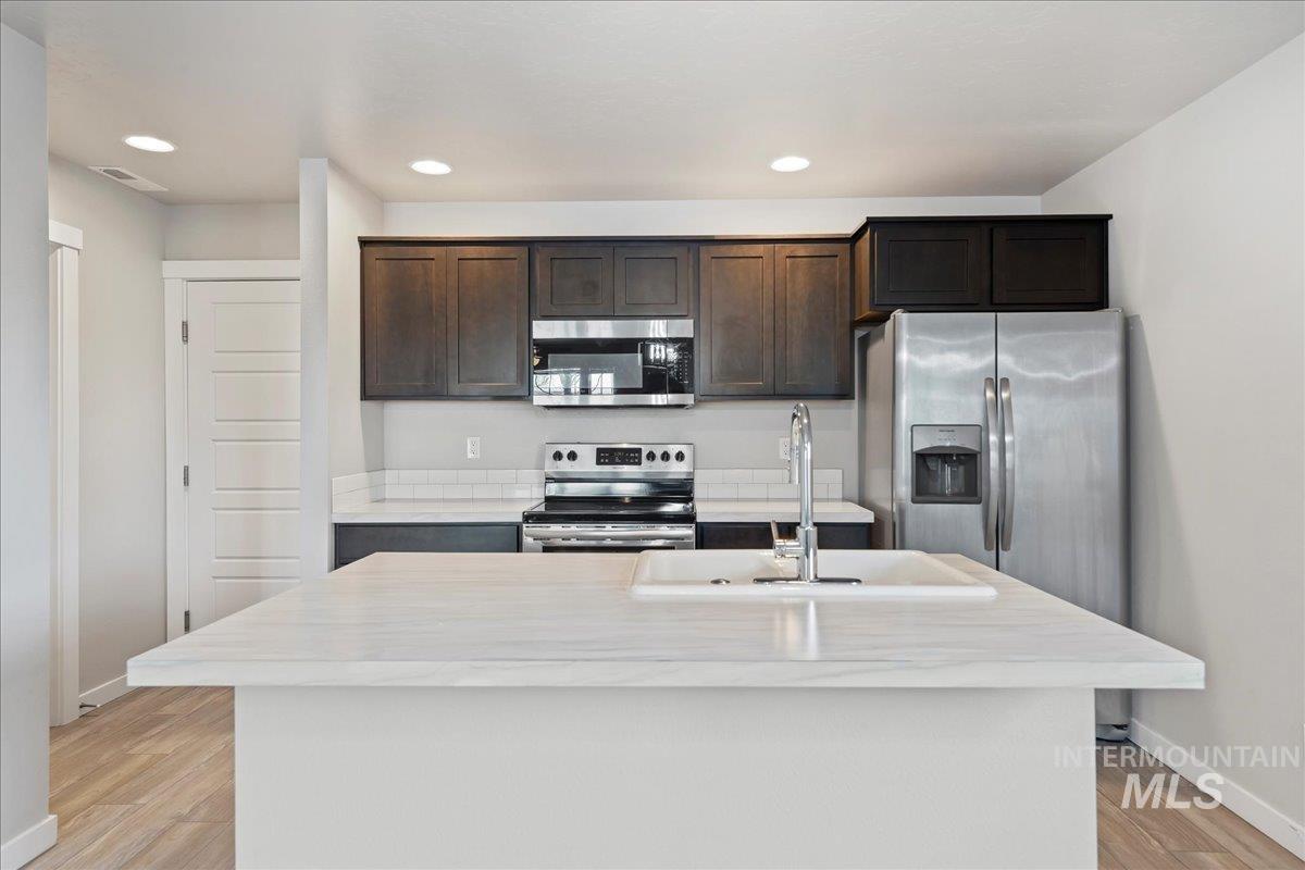 Kitchen featuring appliances with stainless steel finishes, dark brown cabinets, recessed lighting, a center island with sink, and light wood-type flooring