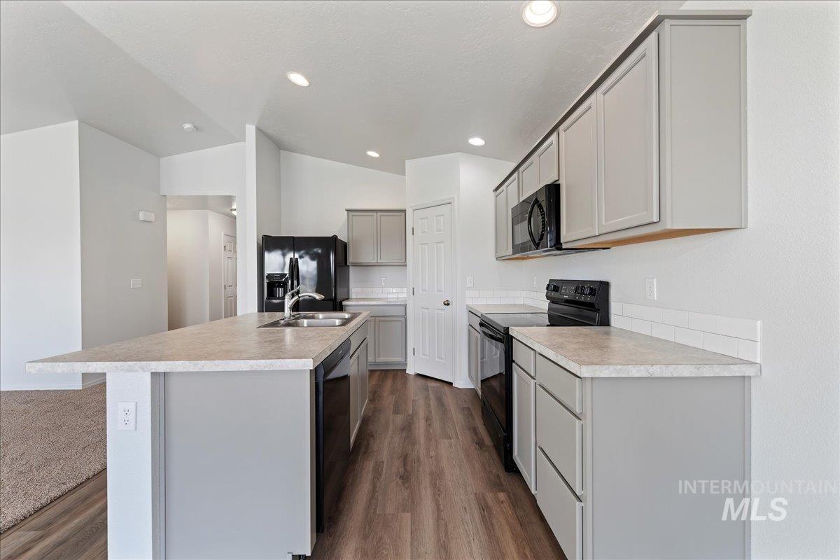 Kitchen with black appliances, vaulted ceiling, light countertops, a center island with sink, and dark wood-style flooring