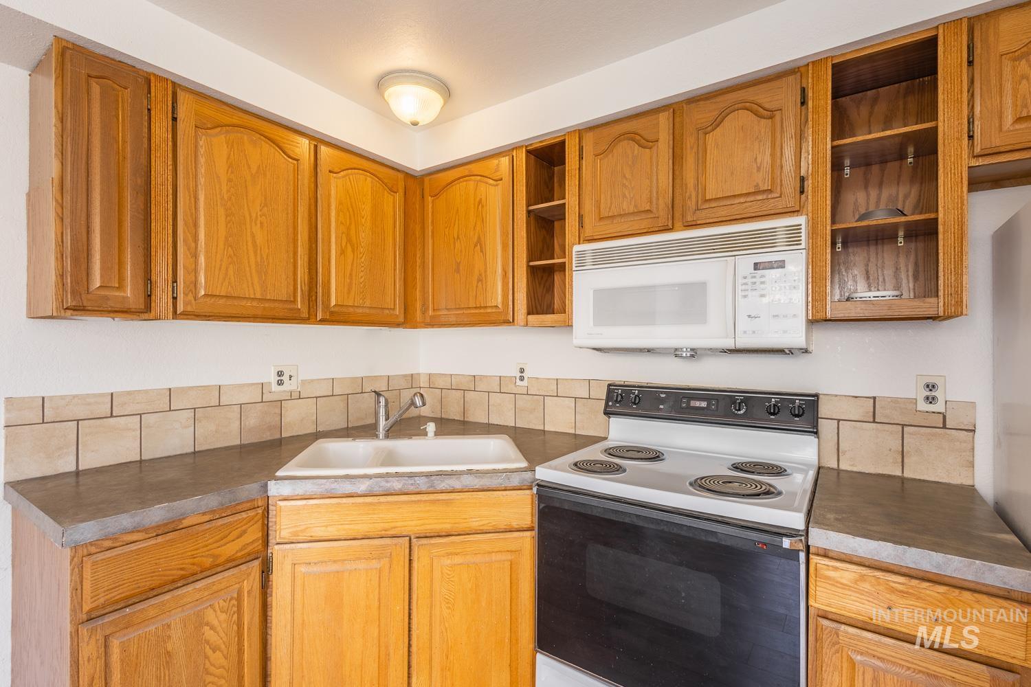 Kitchen with electric stove, open shelves, white microwave, brown cabinetry, and dark countertops