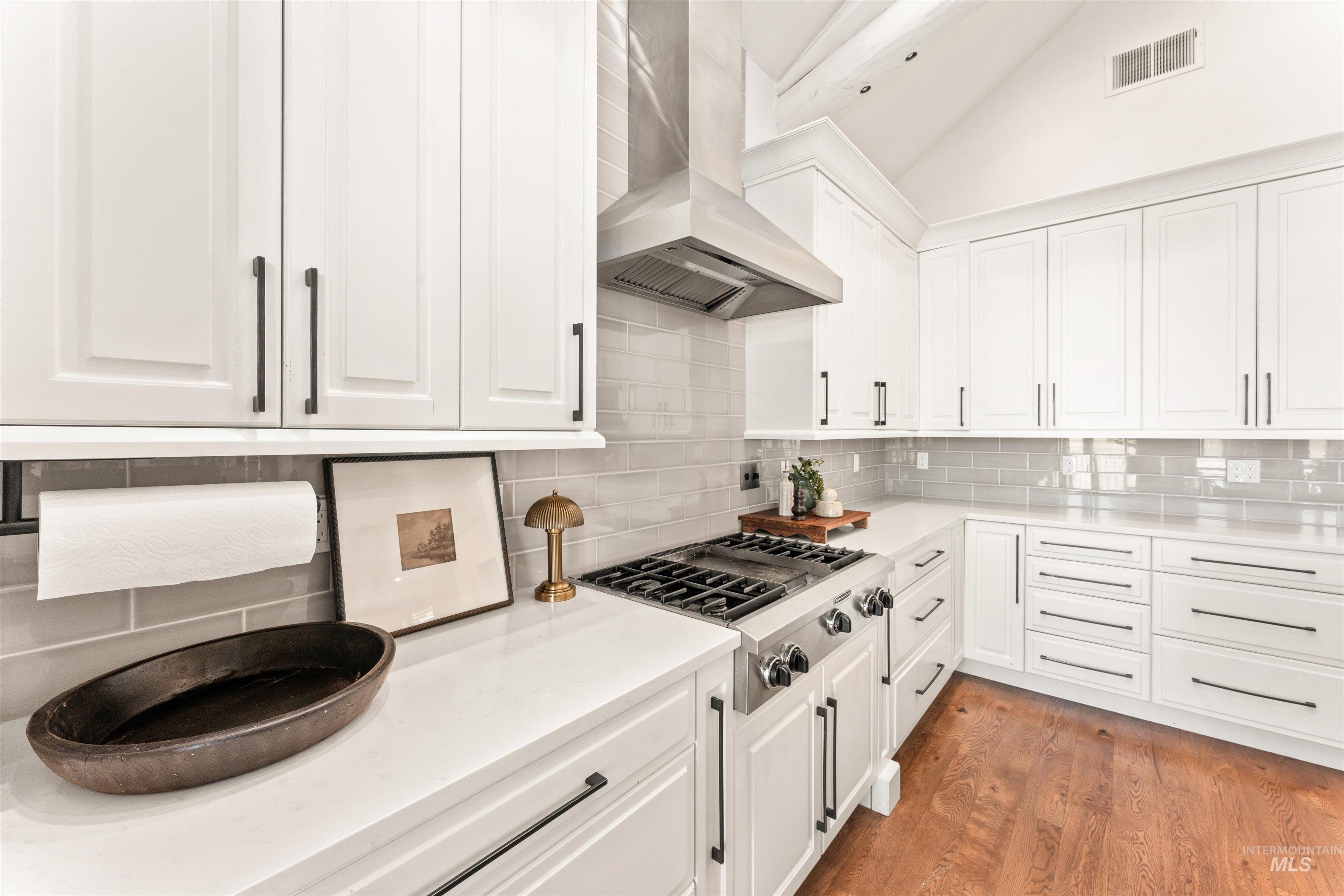 Kitchen featuring white cabinets, wall chimney range hood, stainless steel gas cooktop, backsplash, and vaulted ceiling