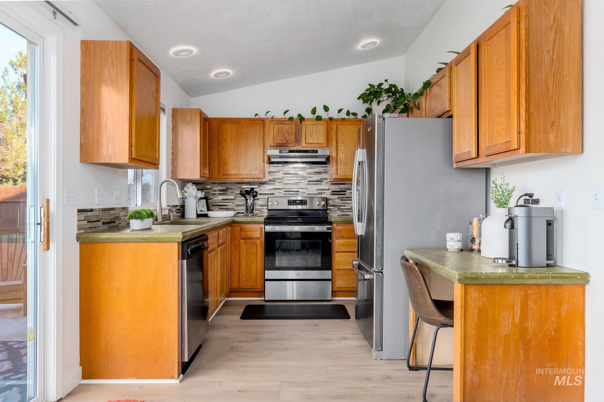 Kitchen featuring stainless steel appliances, vaulted ceiling, brown cabinetry, decorative backsplash, and light countertops