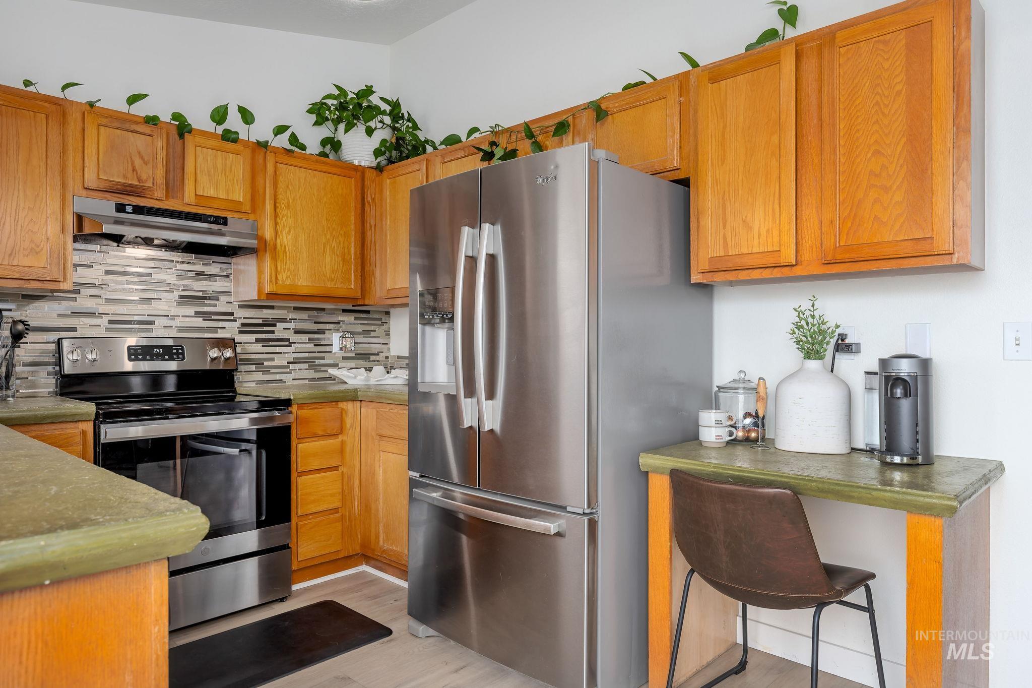 Kitchen with appliances with stainless steel finishes, under cabinet range hood, backsplash, brown cabinets, and dark countertops