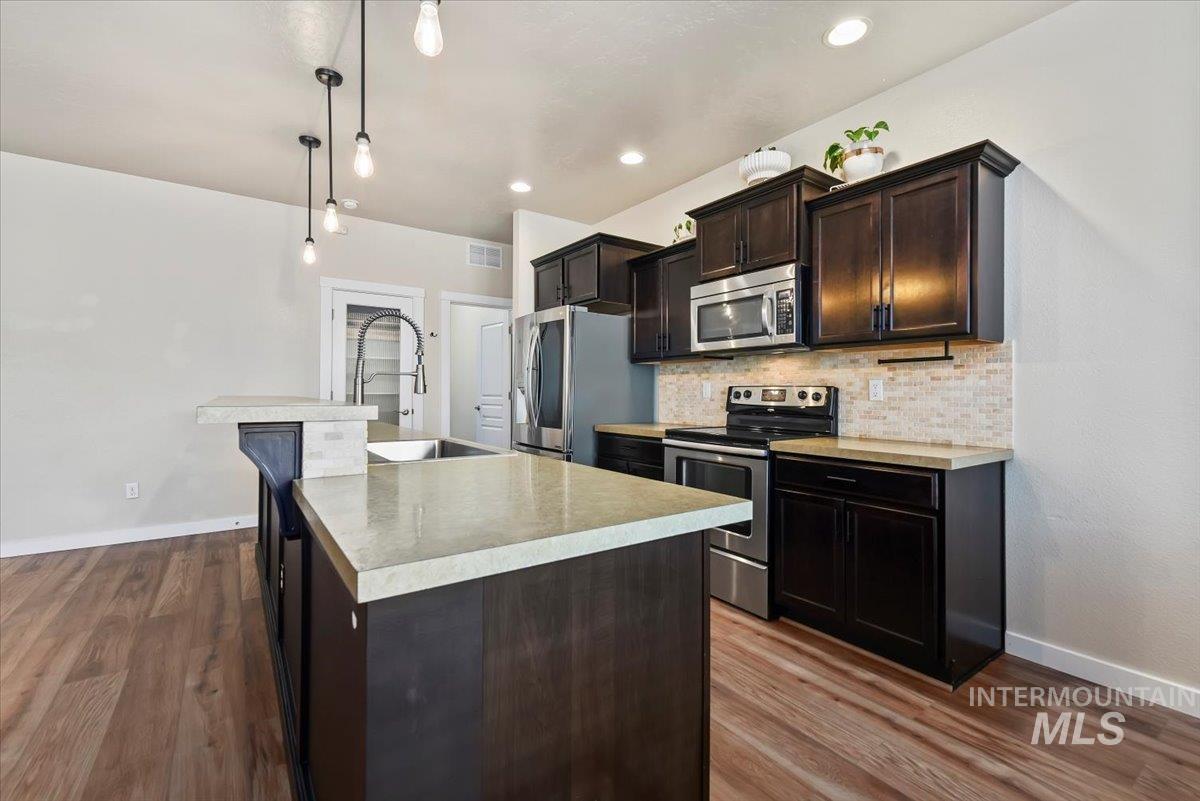 Kitchen featuring stainless steel appliances, tasteful backsplash, decorative light fixtures, dark brown cabinets, and light countertops