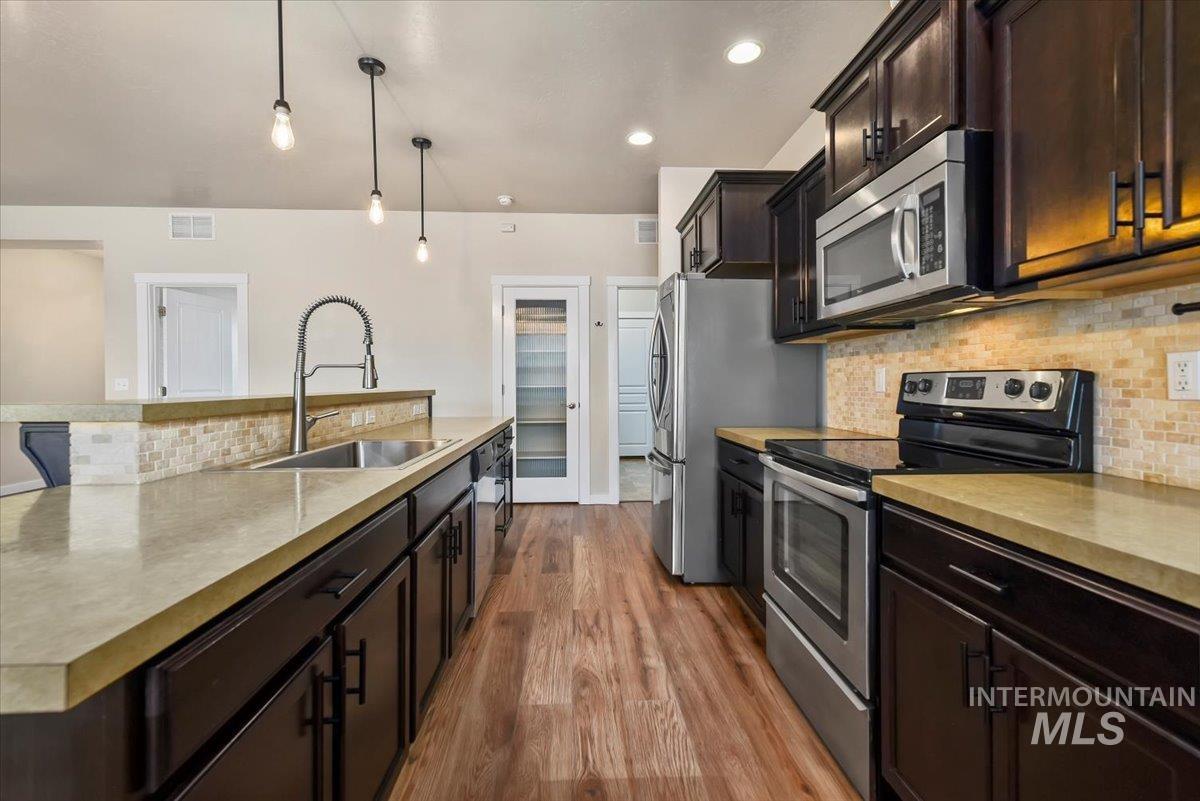 Kitchen featuring decorative backsplash, stainless steel appliances, dark brown cabinetry, a kitchen island with sink, and decorative light fixtures