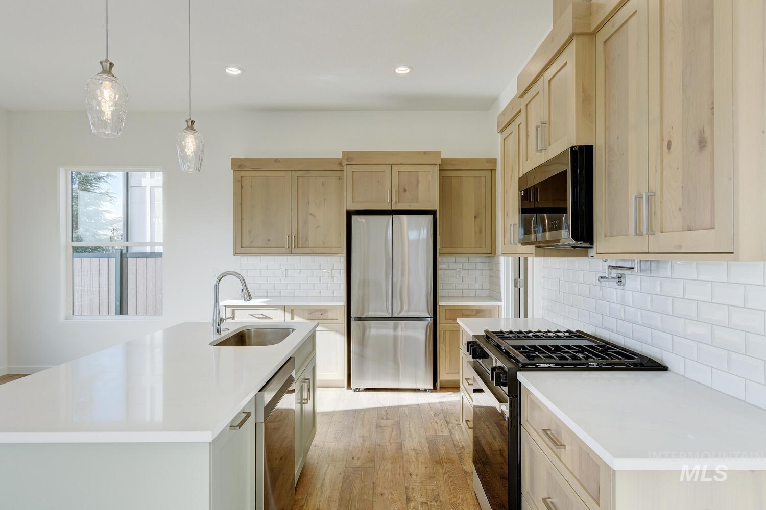 Kitchen featuring appliances with stainless steel finishes, hanging light fixtures, light brown cabinetry, light wood-style flooring, and a center island with sink