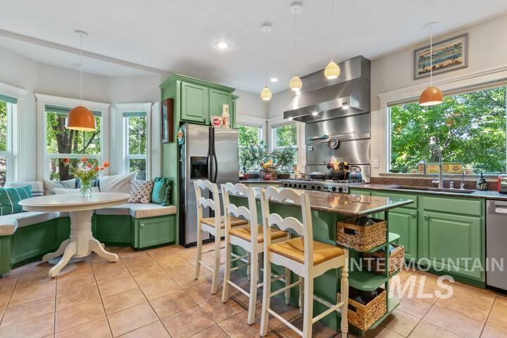 Kitchen featuring green cabinetry, appliances with stainless steel finishes, pendant lighting, a breakfast bar area, and wall chimney exhaust hood
