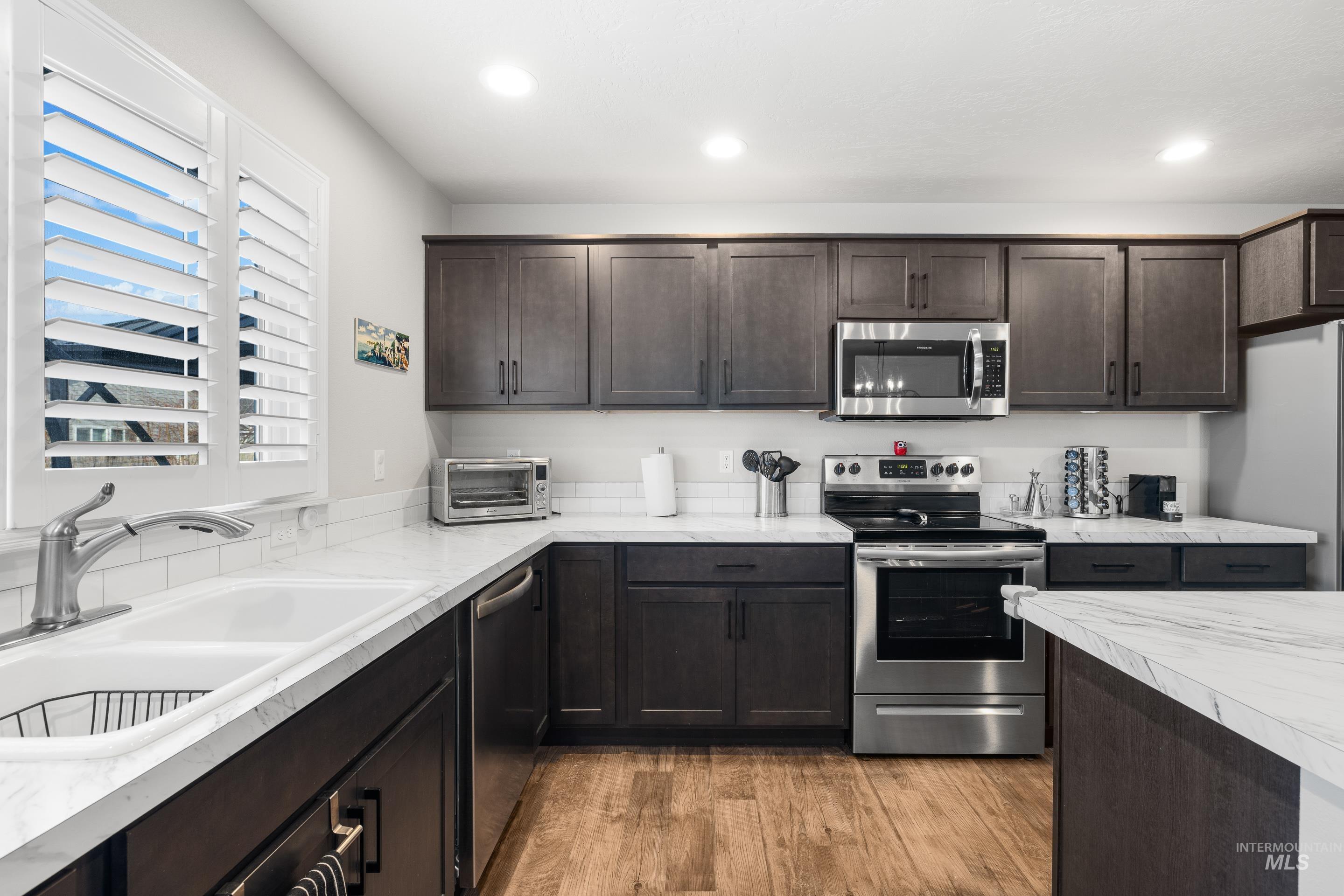 Kitchen with appliances with stainless steel finishes, dark brown cabinetry, light countertops, light wood-type flooring, and recessed lighting