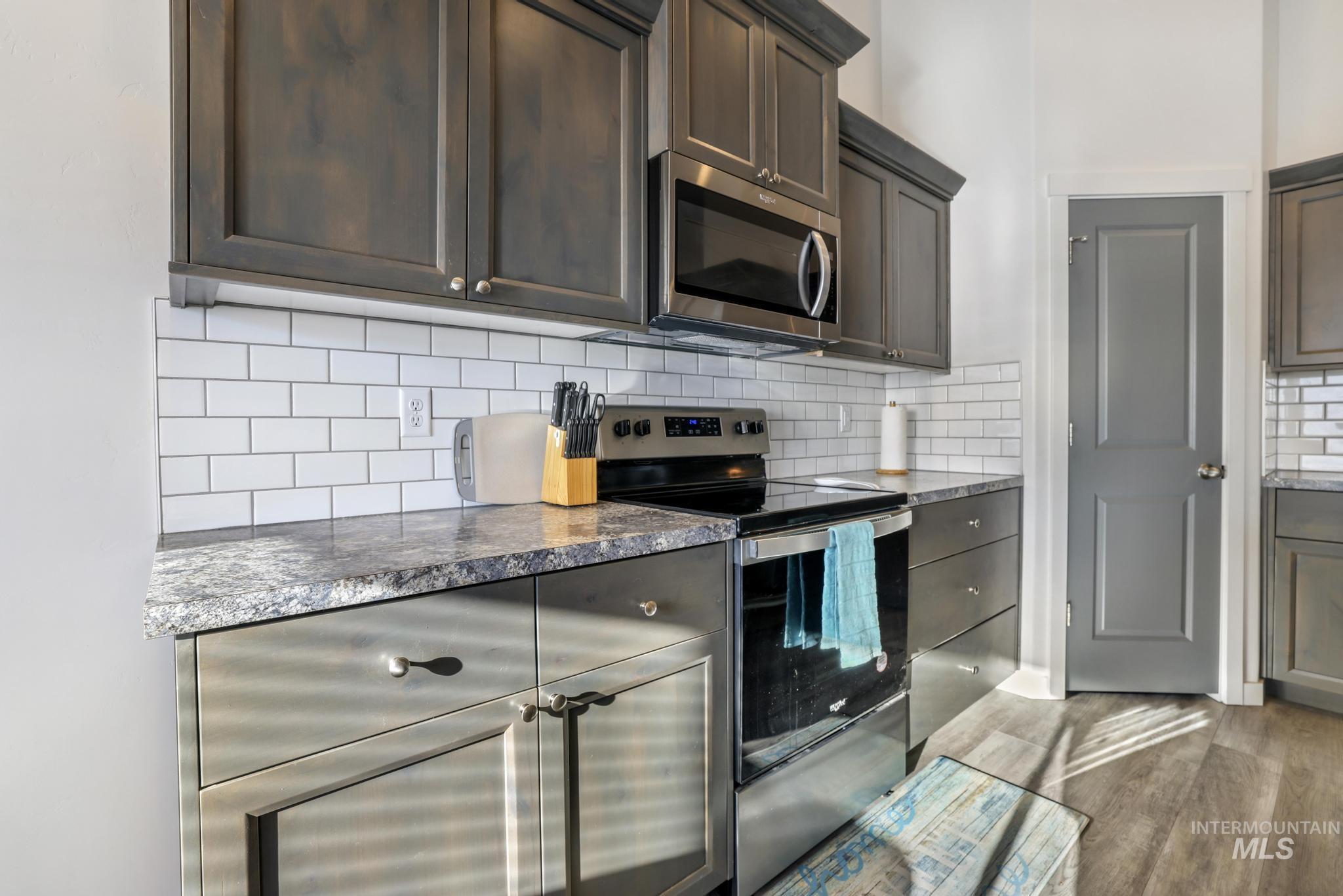 Kitchen with stainless steel appliances, backsplash, light wood finished floors, dark brown cabinetry, and dark stone countertops