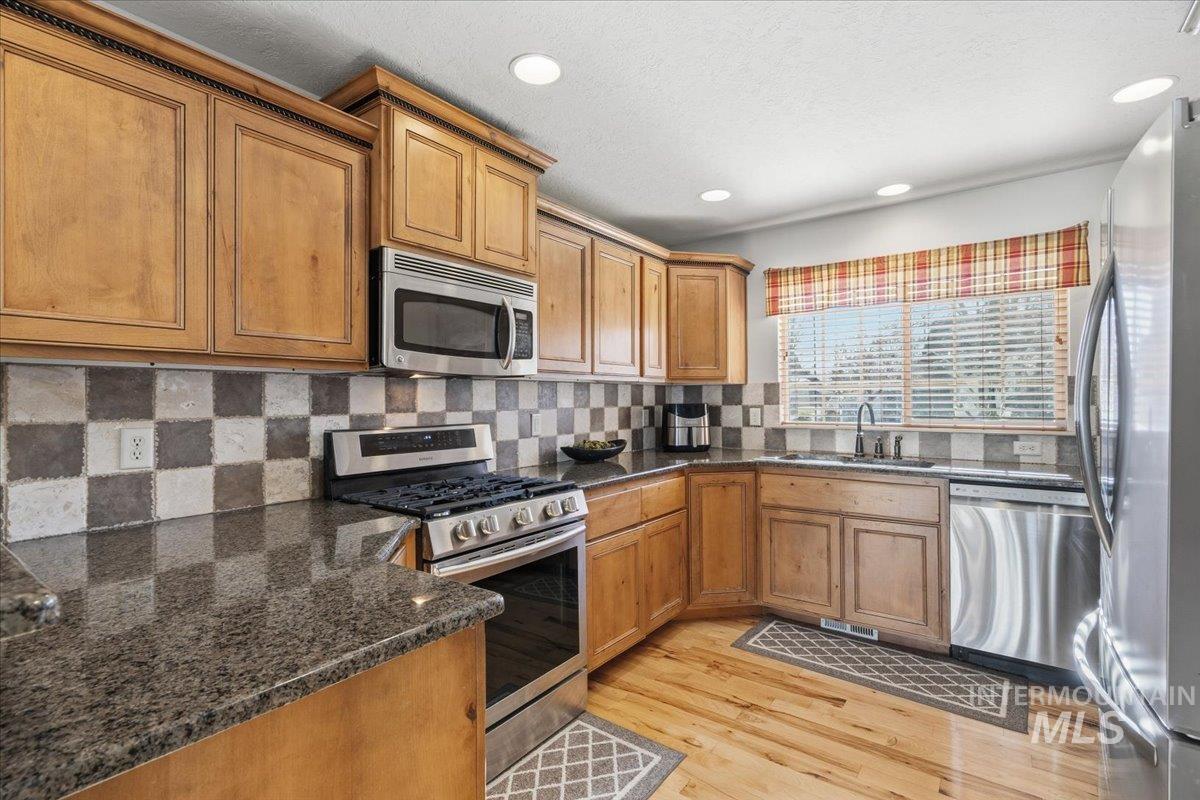 Kitchen featuring stainless steel appliances, brown cabinetry, tasteful backsplash, dark stone countertops, and light wood-style floors