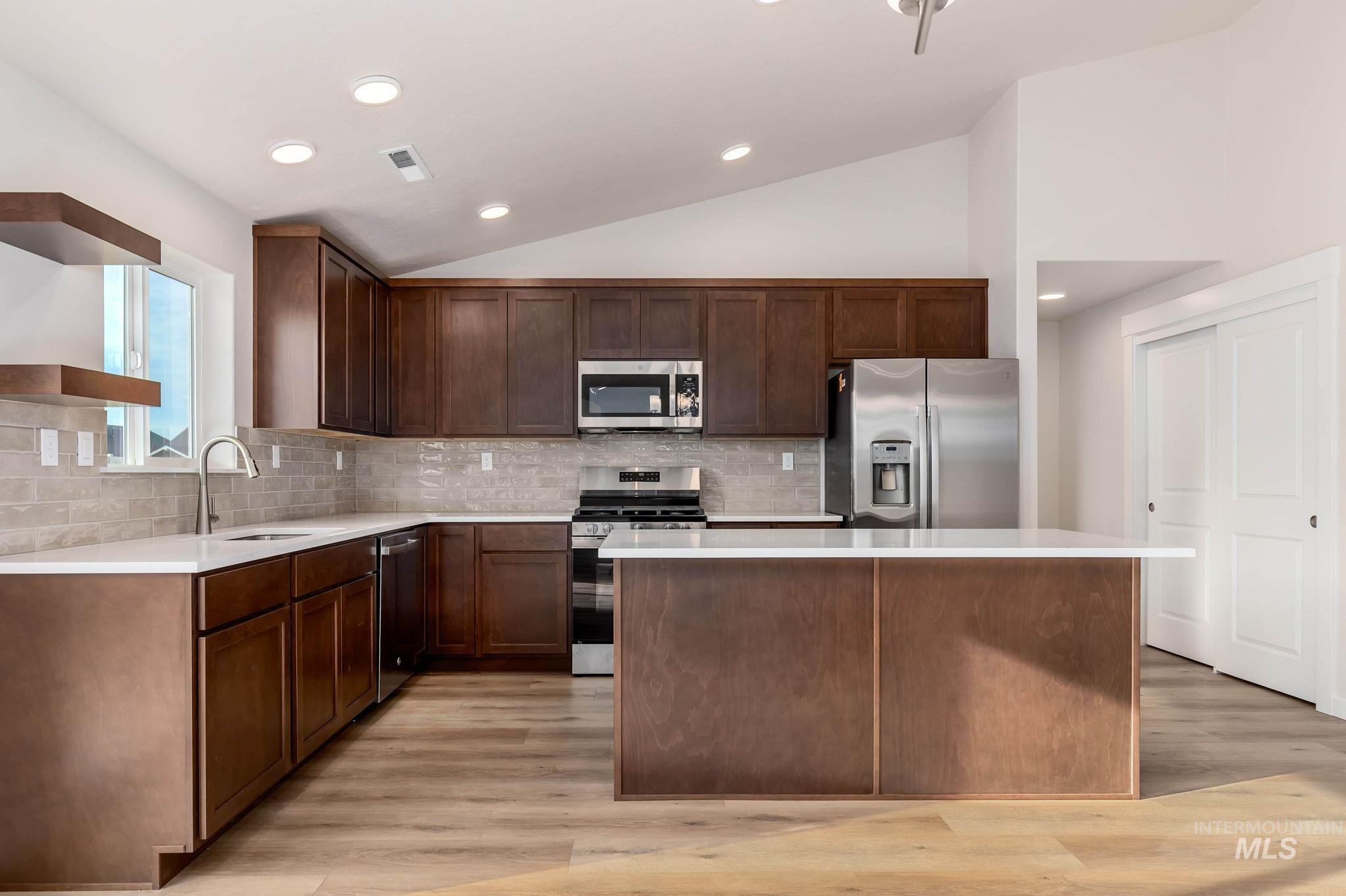 Kitchen featuring lofted ceiling, appliances with stainless steel finishes, light wood finished floors, decorative backsplash, and a kitchen island