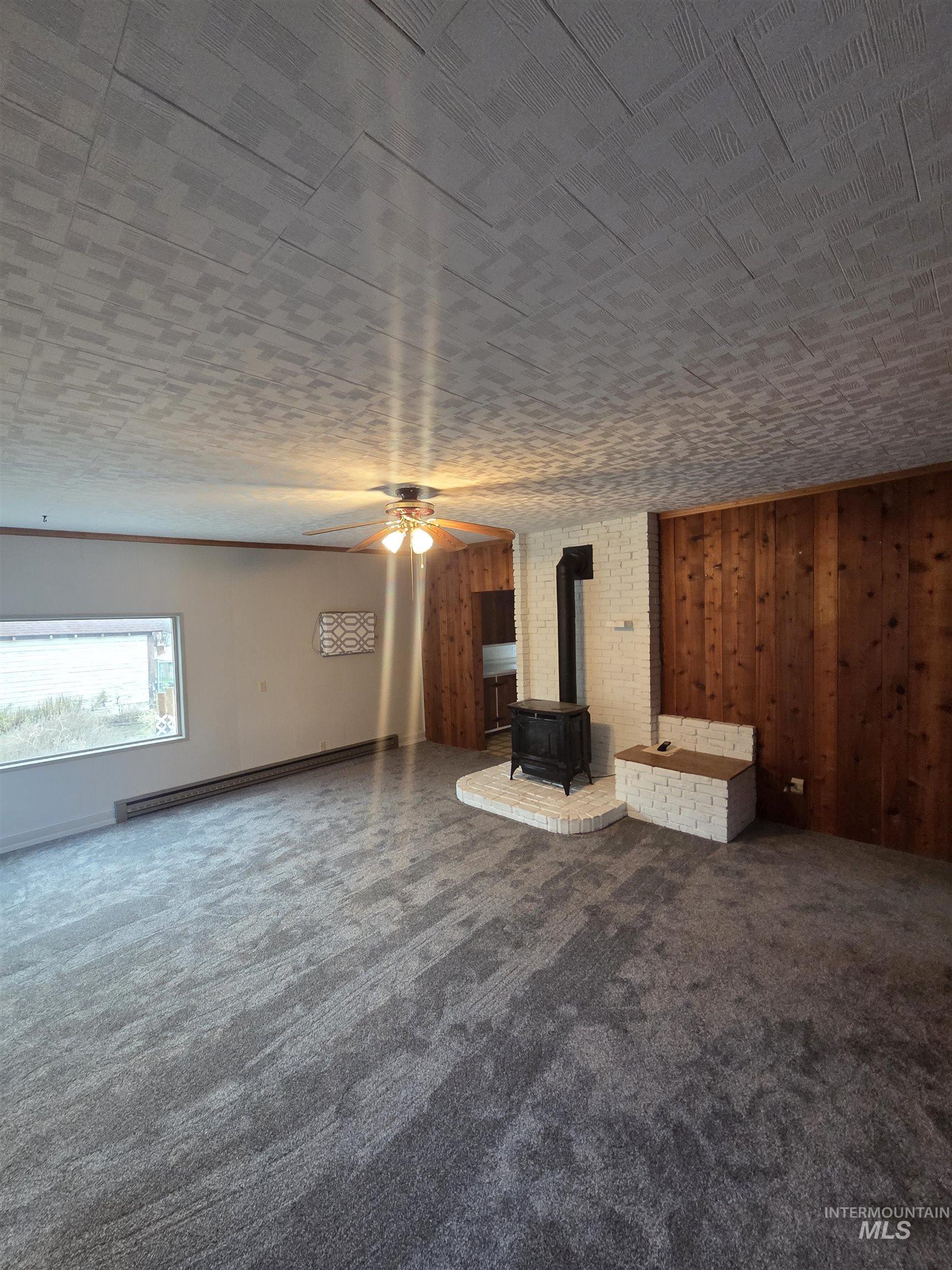 Unfurnished living room featuring a wood stove, a ceiling fan, dark carpet, a baseboard radiator, and wooden walls