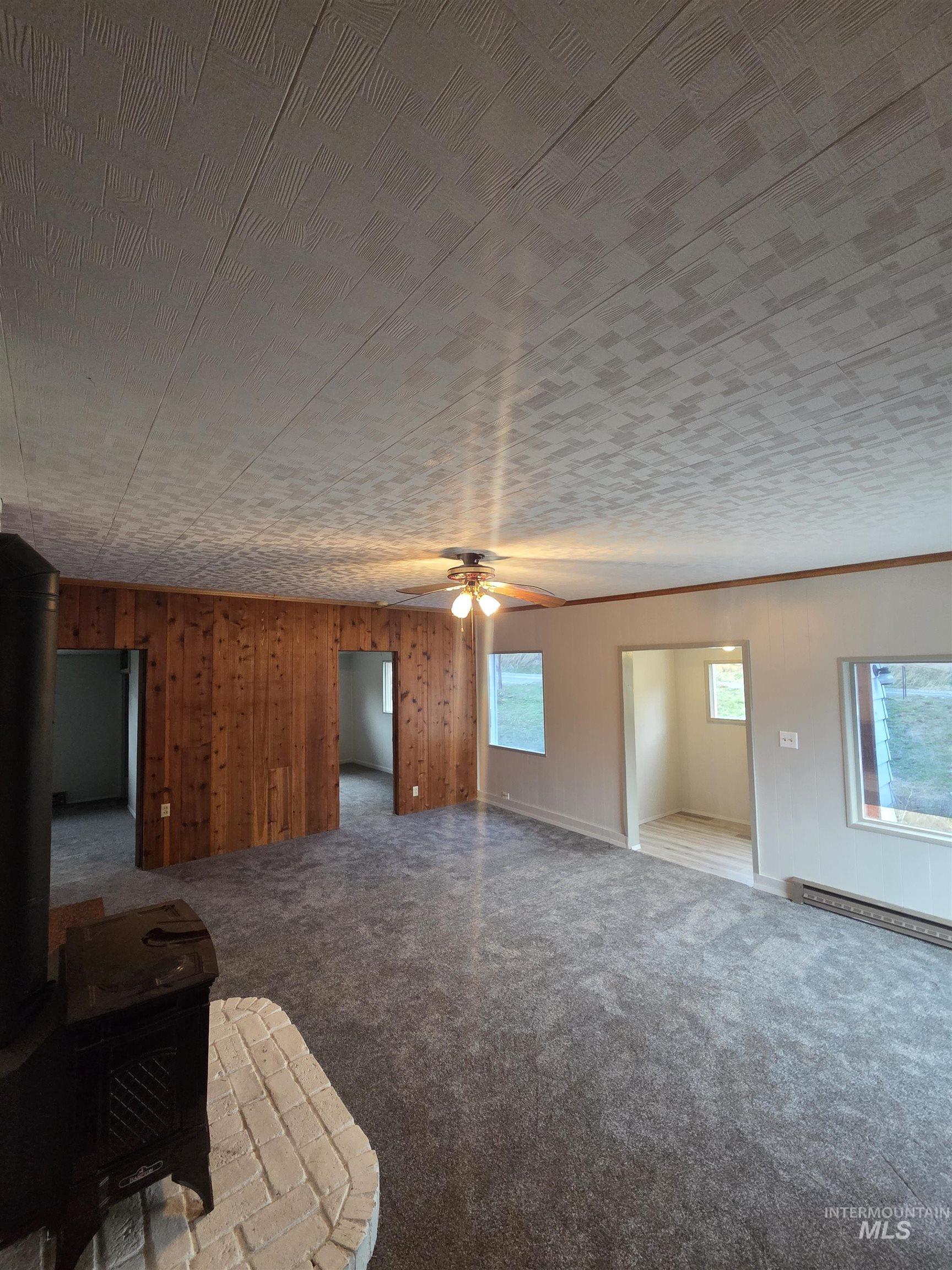 Unfurnished living room featuring wooden walls, a wood stove, carpet, a baseboard heating unit, and ceiling fan