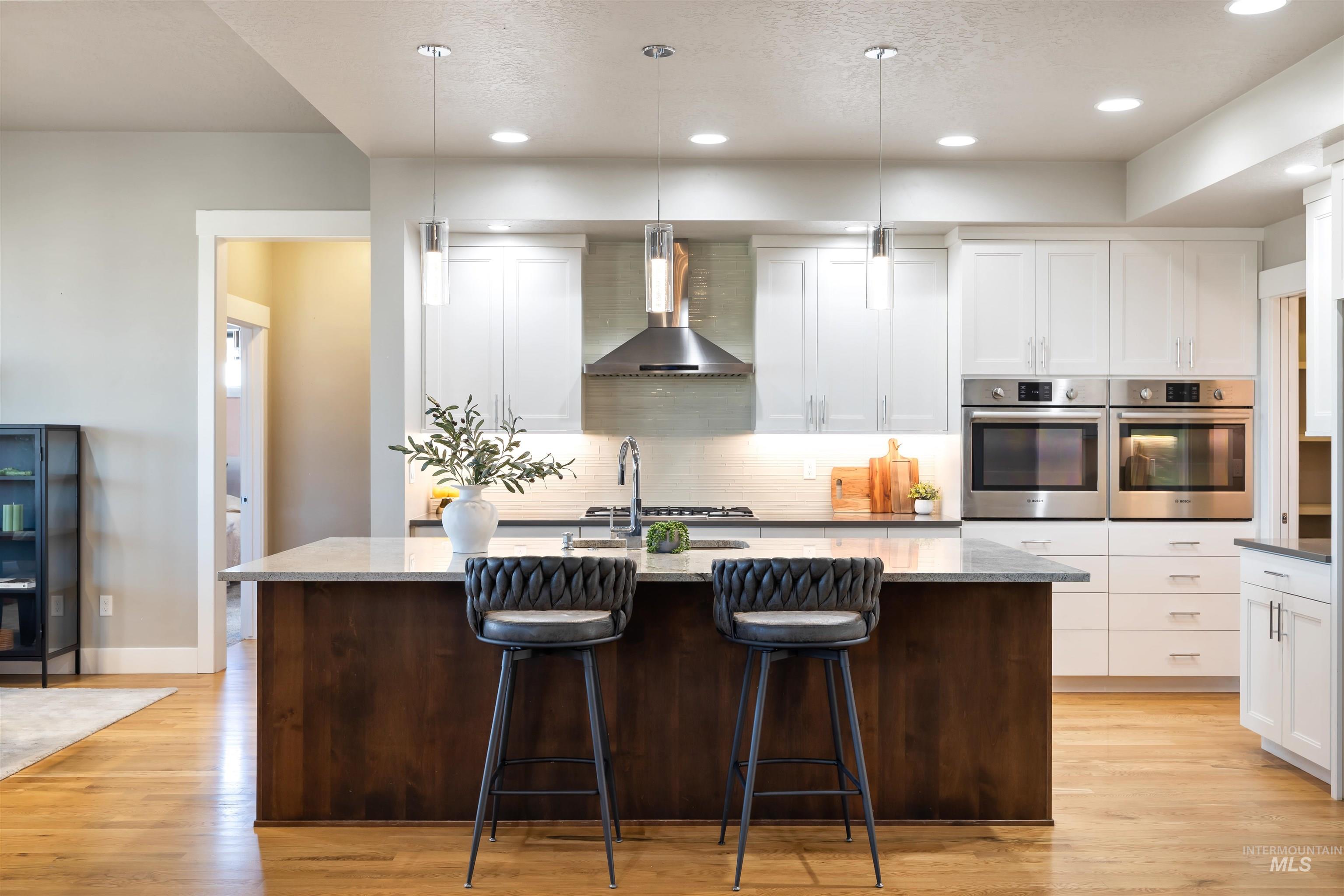 Kitchen with dark brown cabinets, tasteful backsplash, a kitchen island with sink, wall chimney exhaust hood, and white cabinetry