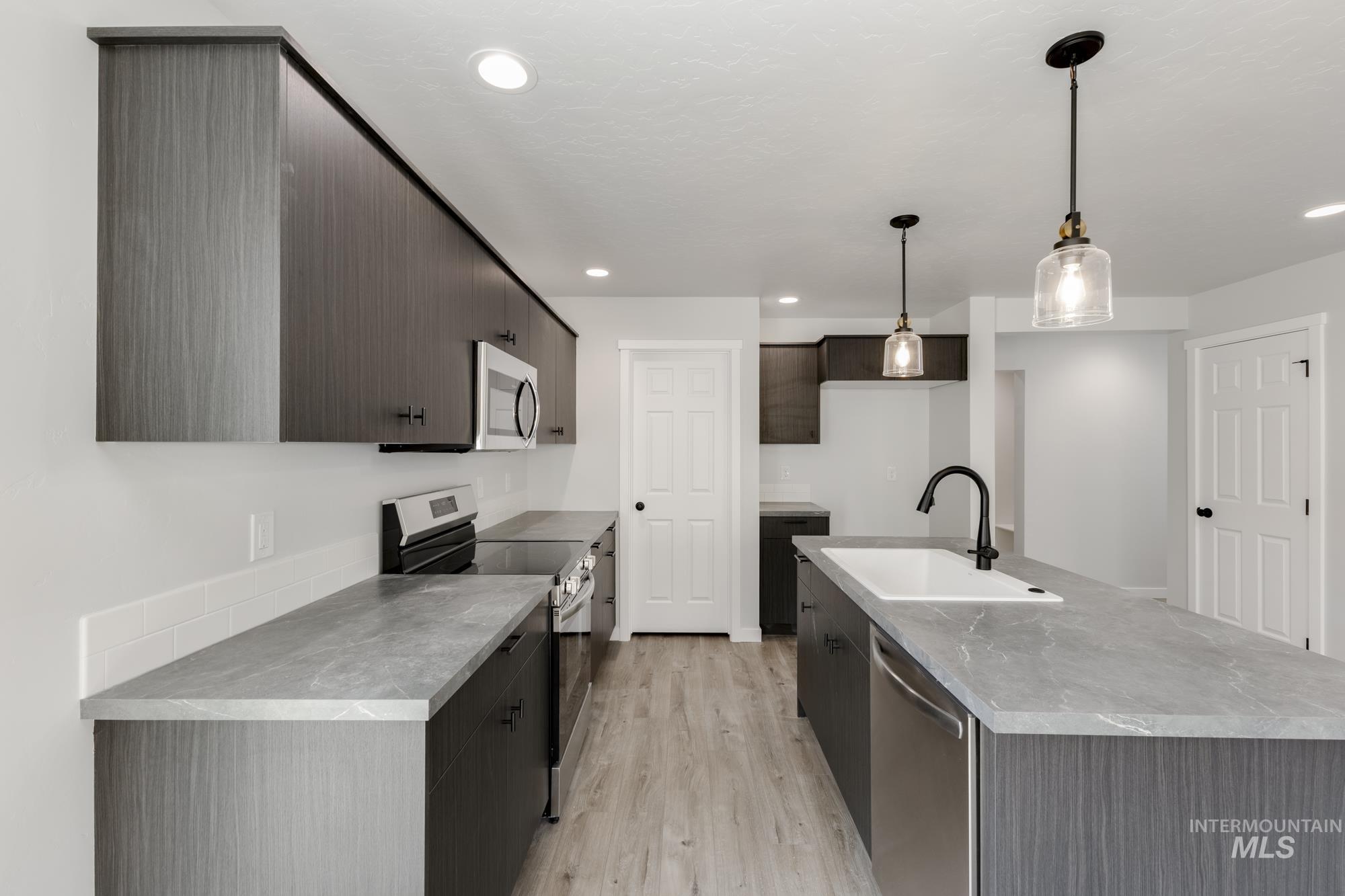 Kitchen with stainless steel appliances, hanging light fixtures, light wood finished floors, a center island with sink, and dark brown cabinets