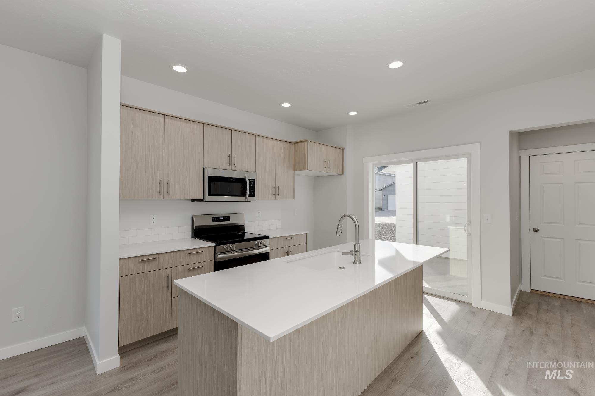 Kitchen with light brown cabinetry, appliances with stainless steel finishes, an island with sink, light wood finished floors, and recessed lighting