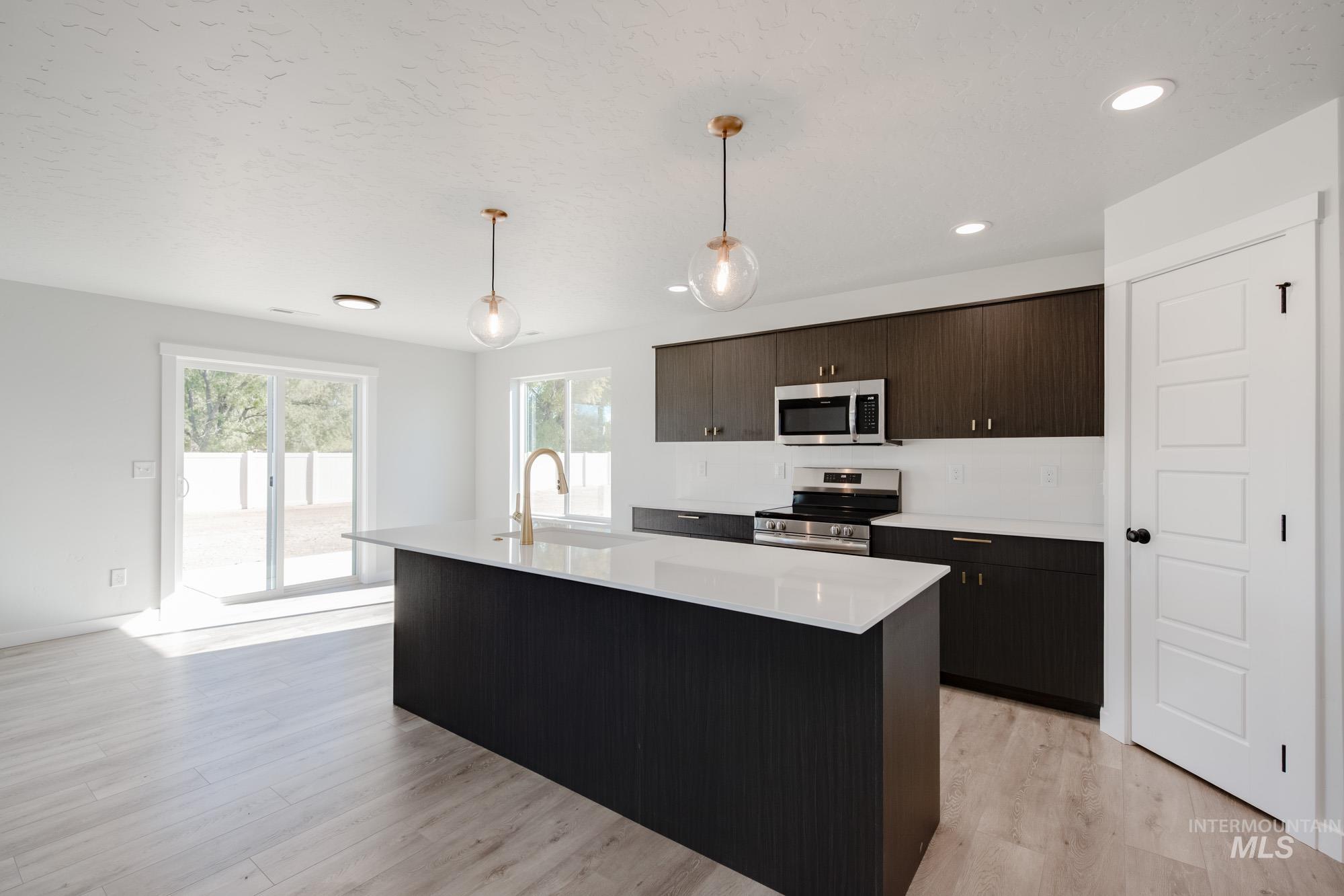 Kitchen with an island with sink, dark brown cabinets, hanging light fixtures, appliances with stainless steel finishes, and light wood finished floors