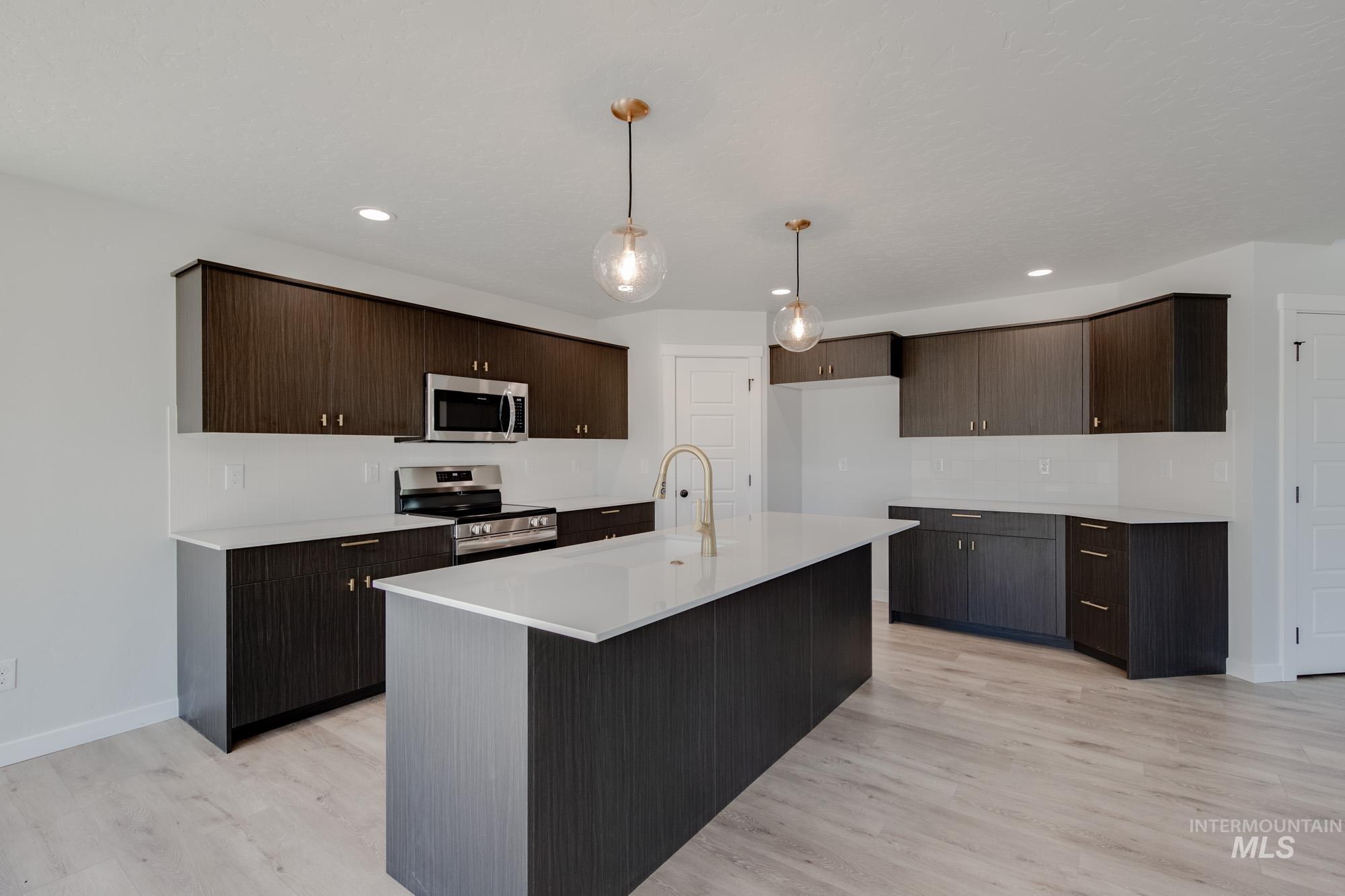 Kitchen with dark brown cabinets, appliances with stainless steel finishes, pendant lighting, light wood-type flooring, and a kitchen island with sink
