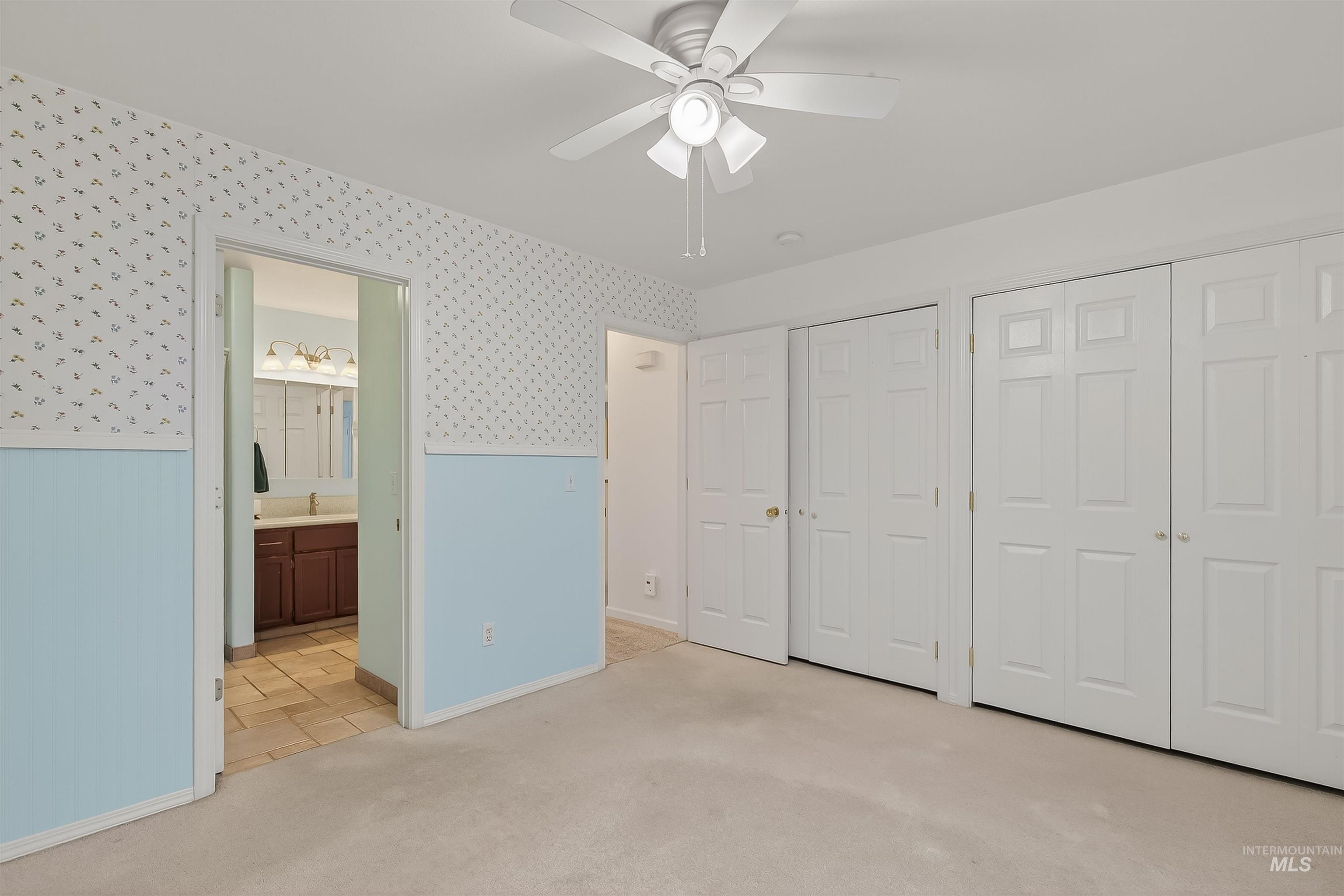 Unfurnished bedroom with wallpapered walls, multiple closets, light colored carpet, wainscoting, and ceiling fan