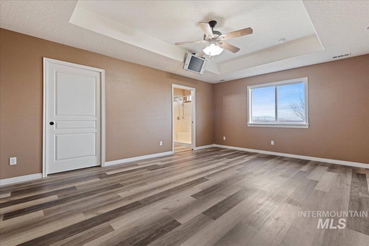 Primary bedroom featuring a tray ceiling, light wood finished floors, a ceiling fan, ensuite bath, and a textured ceiling