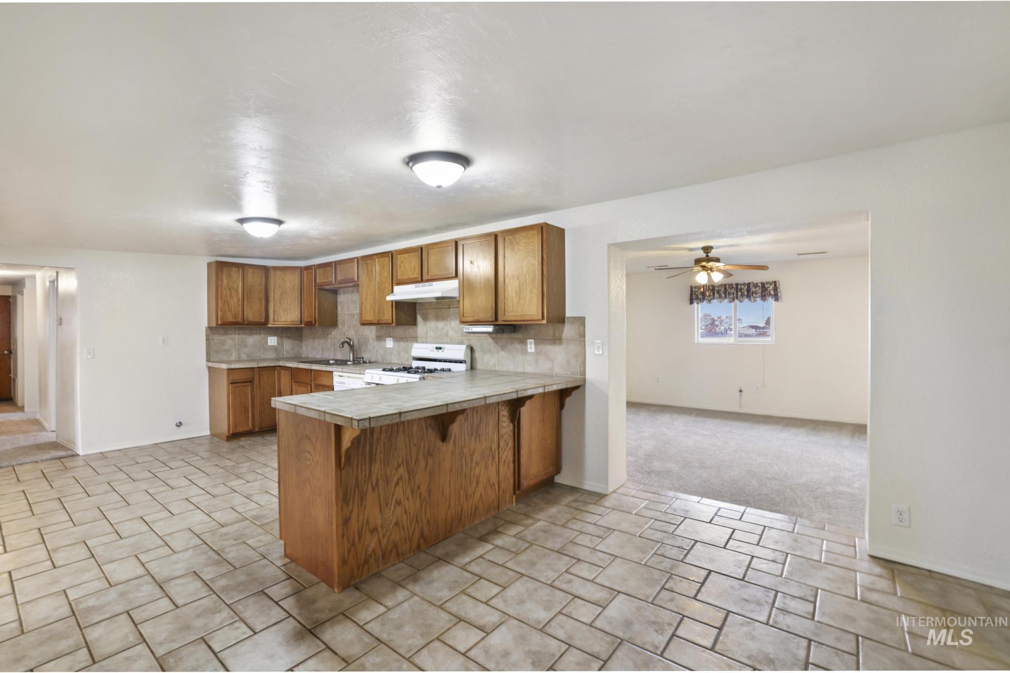 Kitchen featuring brown cabinetry, tile counters, gas range gas stove, a ceiling fan, and open floor plan