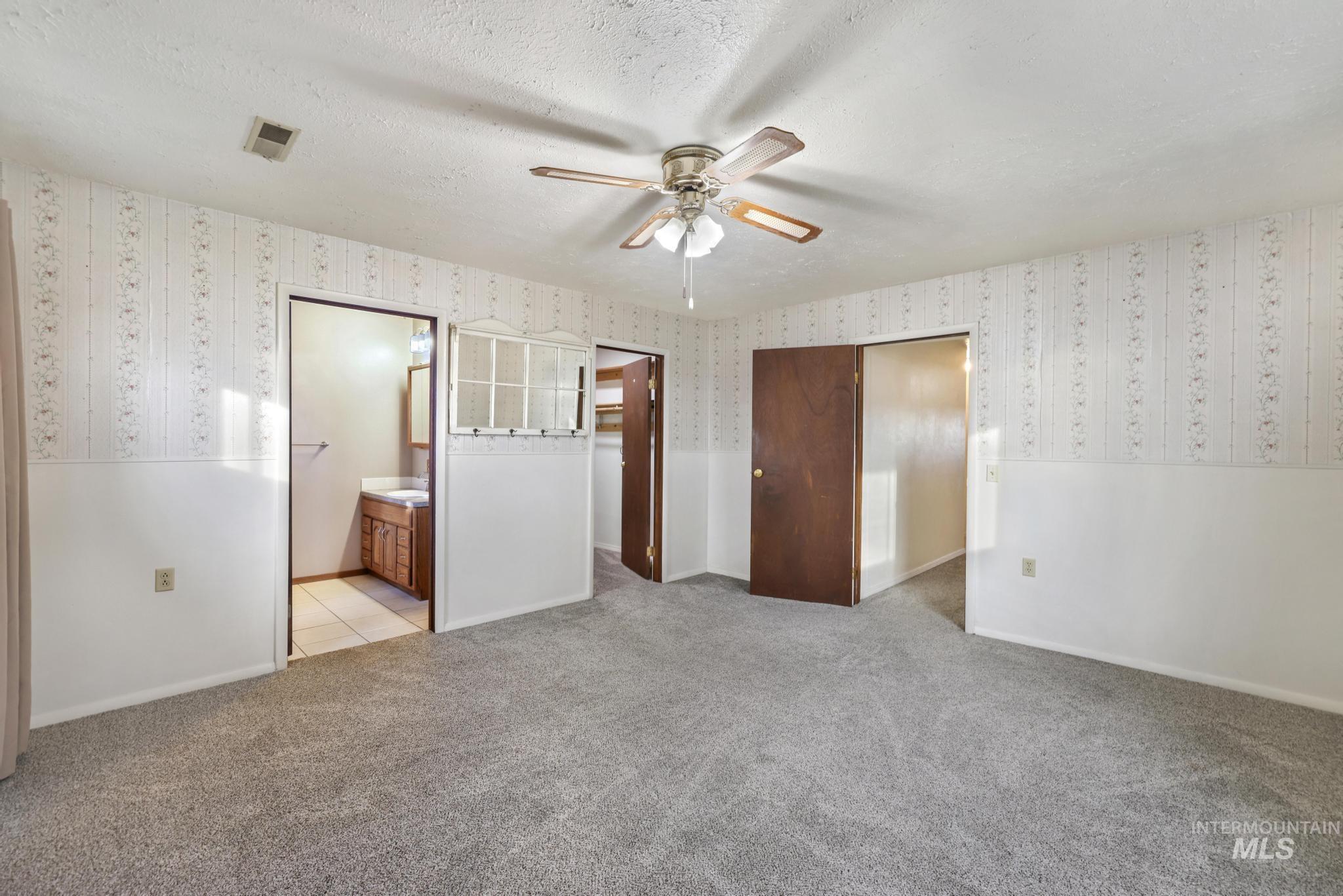 Unfurnished bedroom with light colored carpet, a ceiling fan, a textured ceiling, connected bathroom, and wallpapered walls