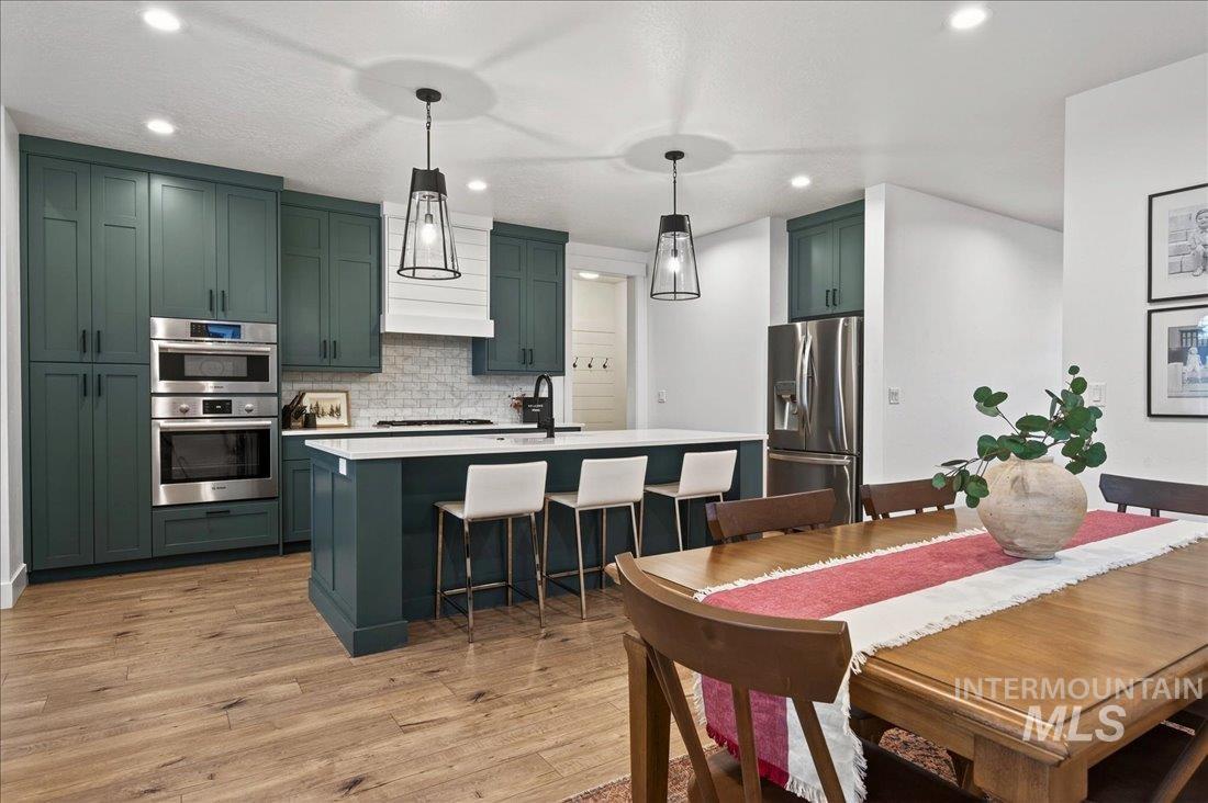 Kitchen with green cabinetry, decorative light fixtures, an island with sink, appliances with stainless steel finishes, and light wood-style flooring