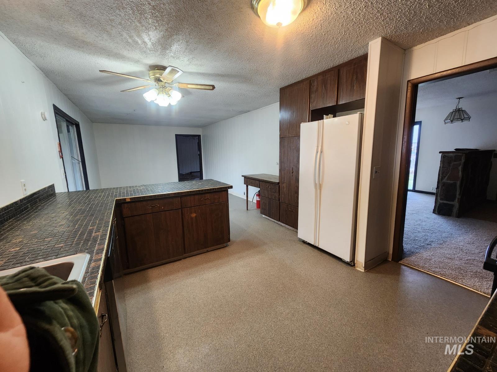 Kitchen with dark brown cabinets, dark countertops, freestanding refrigerator, a ceiling fan, and a textured ceiling