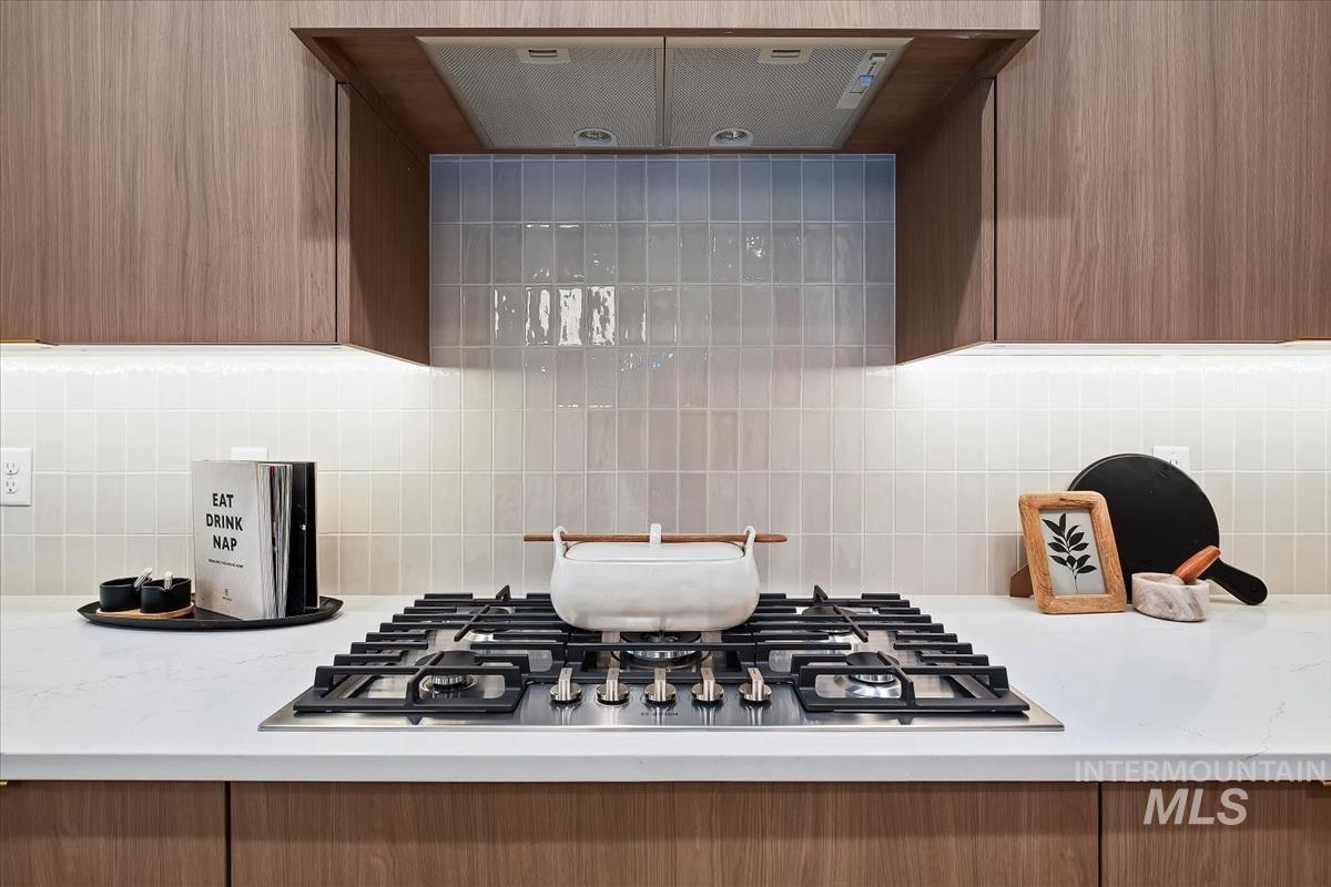 Kitchen view of extractor fan, stainless steel gas cooktop, tasteful backsplash, and light stone counters