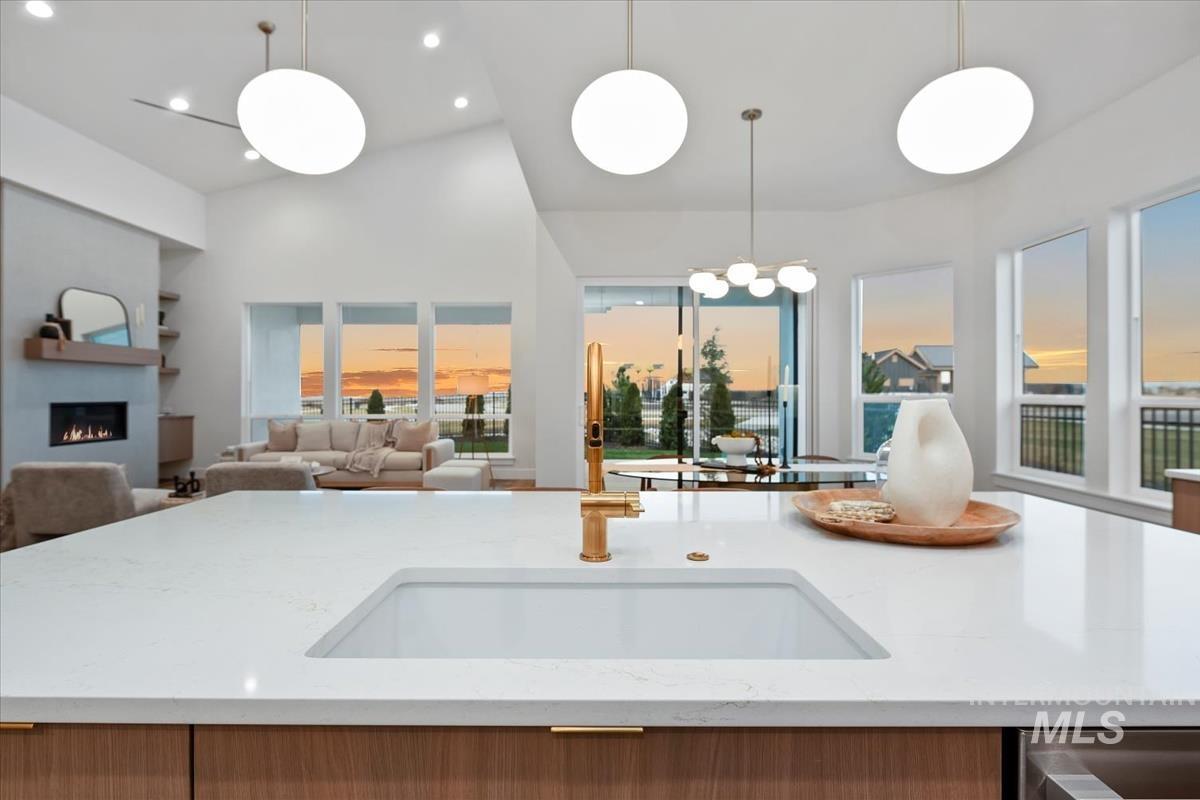 Kitchen with light stone countertops, a warm lit fireplace, an island with sink, decorative light fixtures, and lofted ceiling