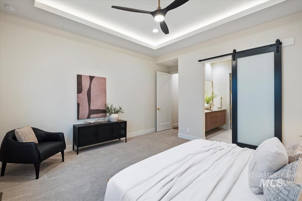 Bedroom with a barn door, light colored carpet, ceiling fan, a raised ceiling, and ensuite bath