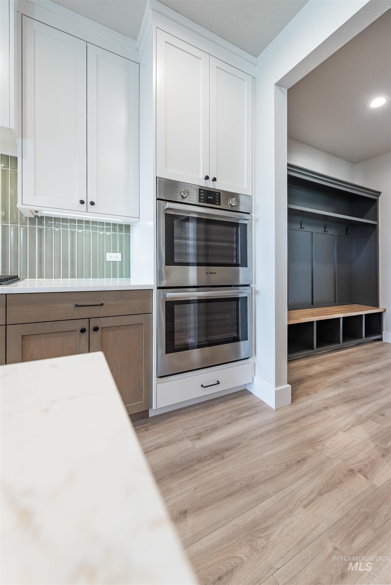 Kitchen featuring stainless steel double oven, white cabinets, brown cabinetry, light wood-style floors, and backsplash