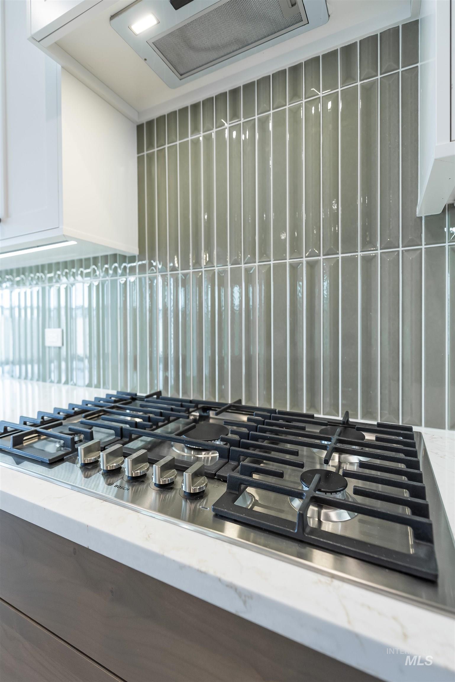 Kitchen view of stainless steel gas stovetop, tasteful backsplash, light stone counters, and white cabinetry