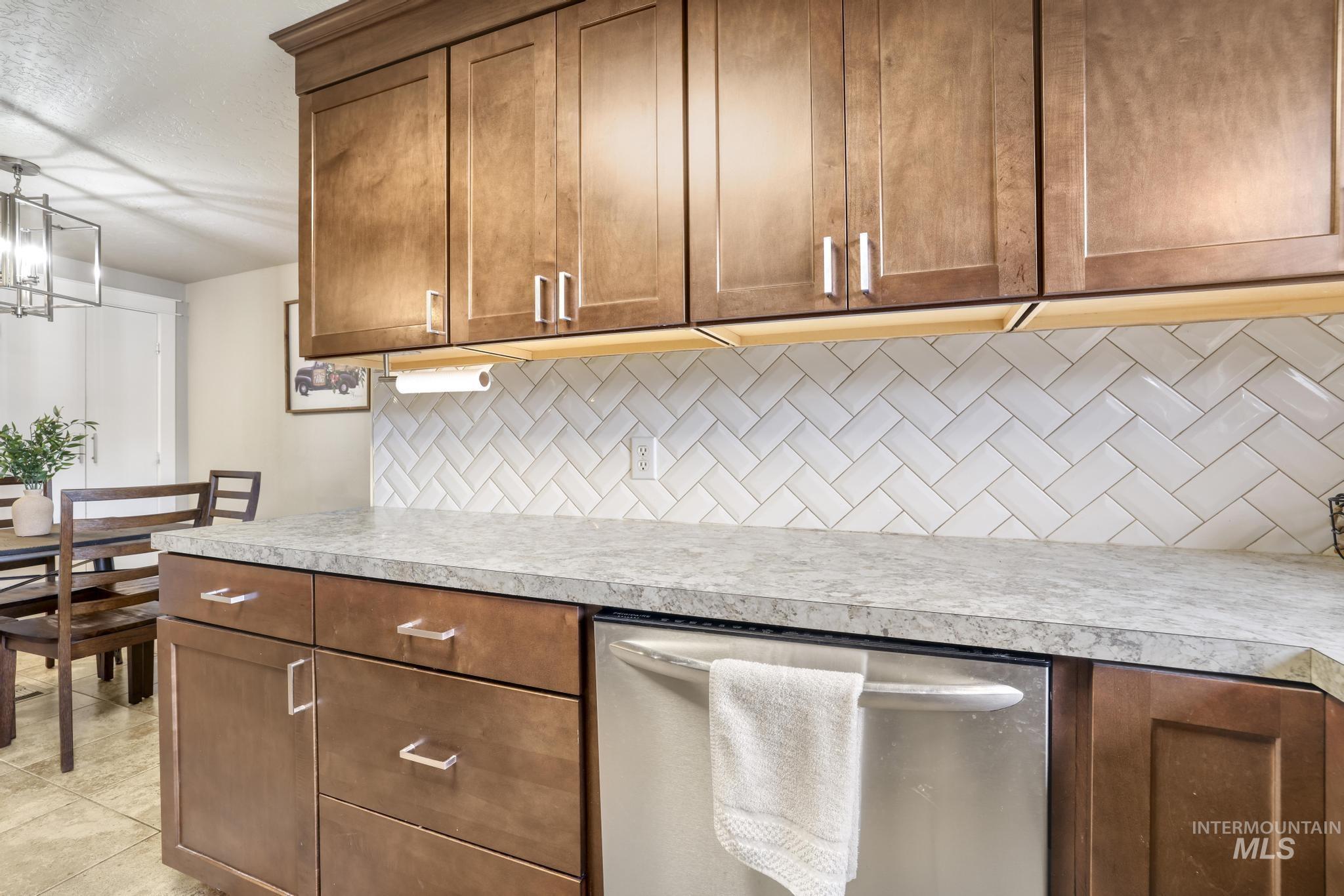 Kitchen featuring stainless steel dishwasher, brown cabinetry, light countertops, and hanging light fixtures