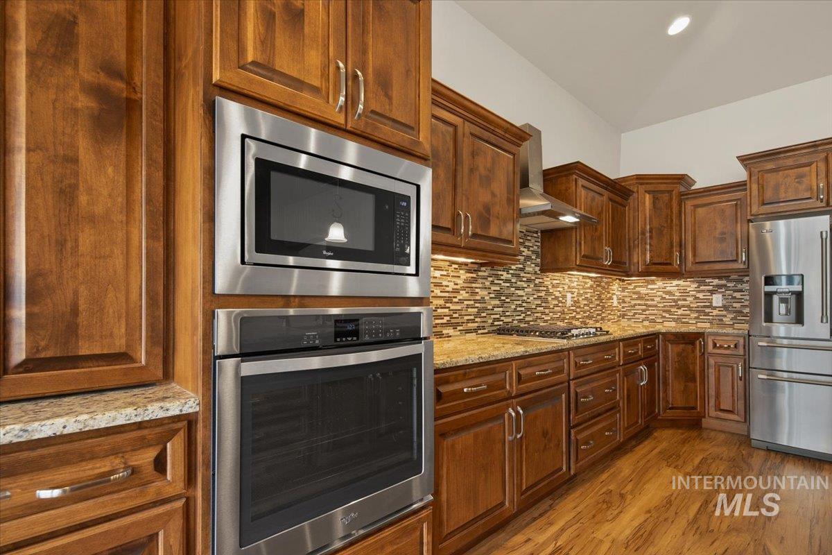 Kitchen with appliances with stainless steel finishes, wall chimney exhaust hood, light stone counters, dark wood finished floors, and tasteful backsplash