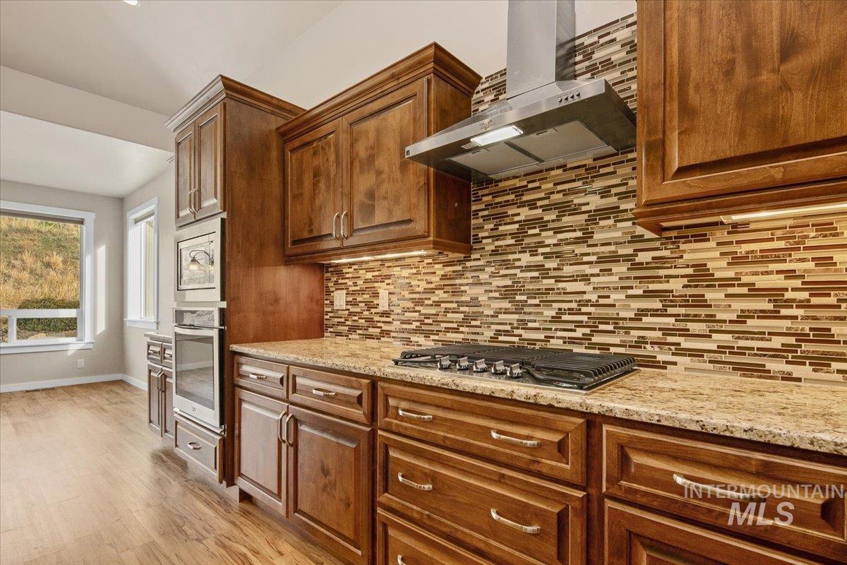 Kitchen featuring wall chimney range hood, light stone countertops, stainless steel appliances, light wood-style flooring, and alder cabinetry